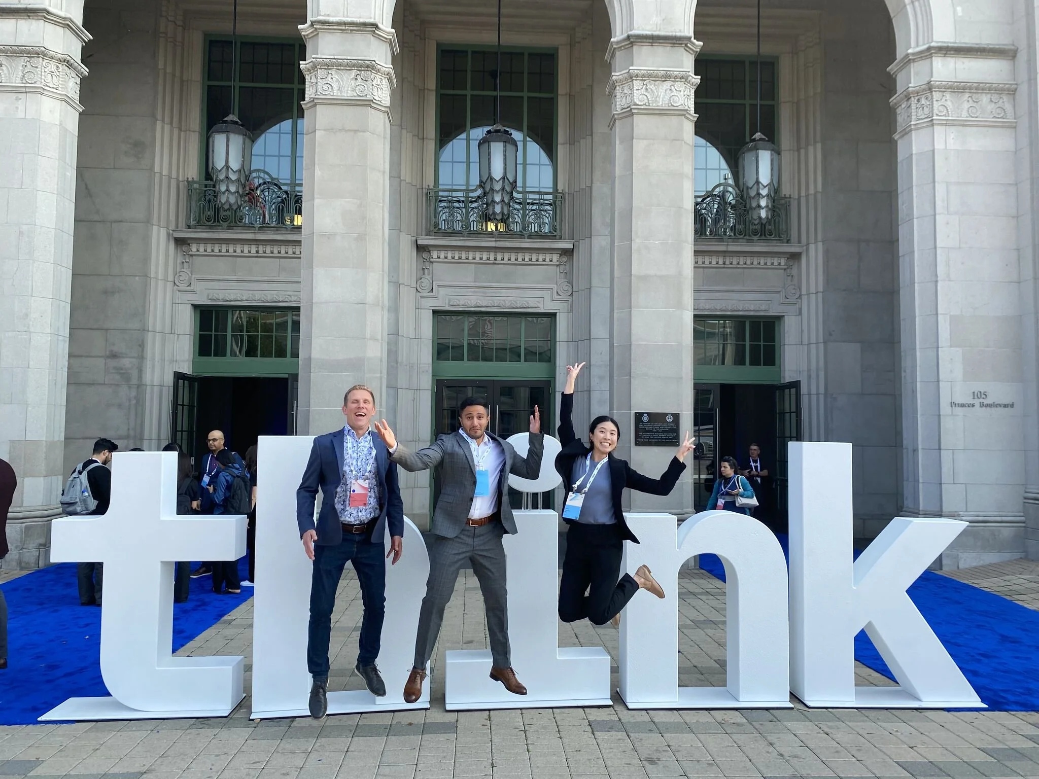 Three people are posing and jumping in front of large white letters that spell 'Eureka' on a blue carpet outside a historic building with large columns and ornate architectural details.