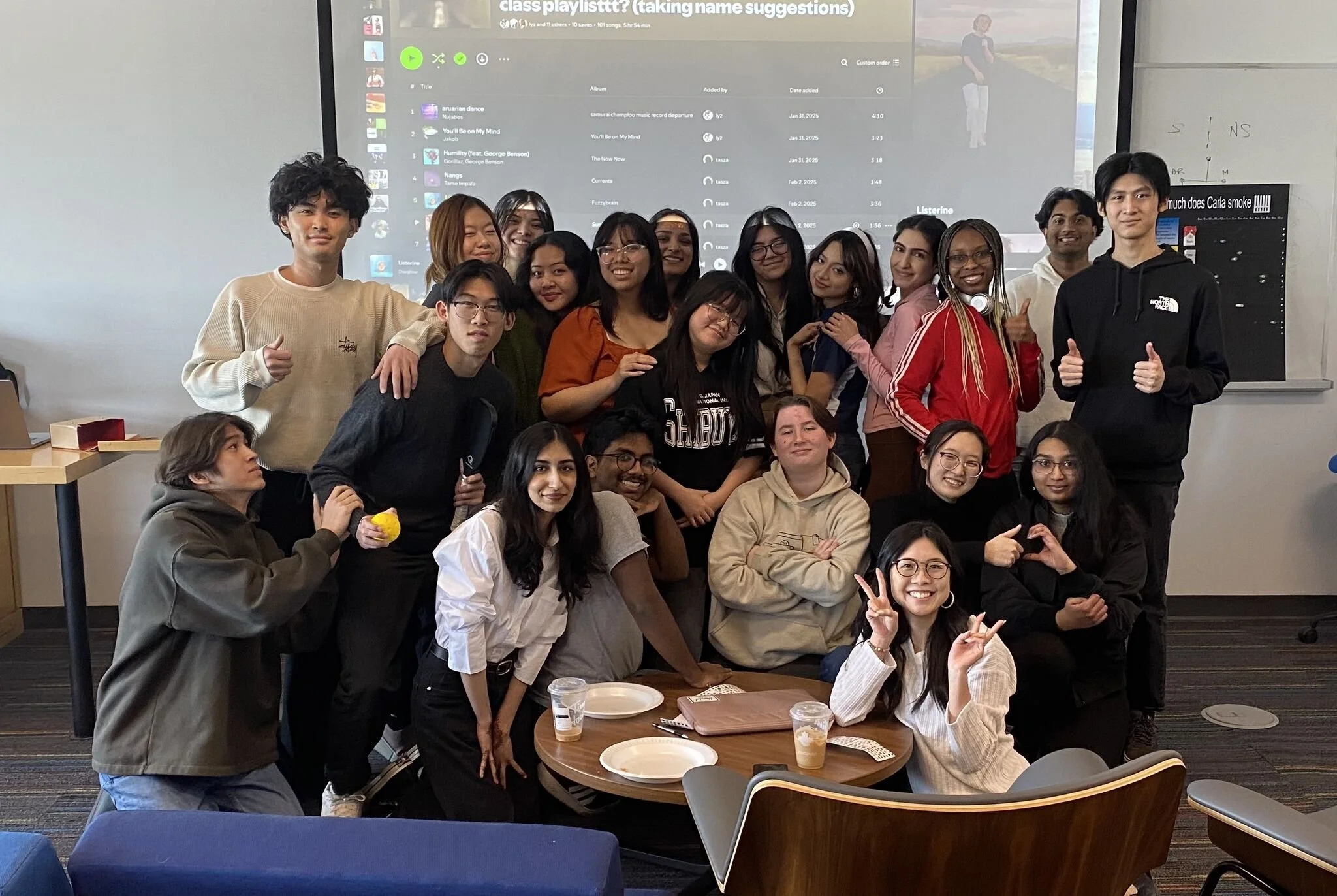 A diverse group of young people posing together in a classroom, some making peace signs or thumbs up. A large monitor behind them displays a playlist on a music streaming platform. There are tables and chairs with empty plates and drinks.