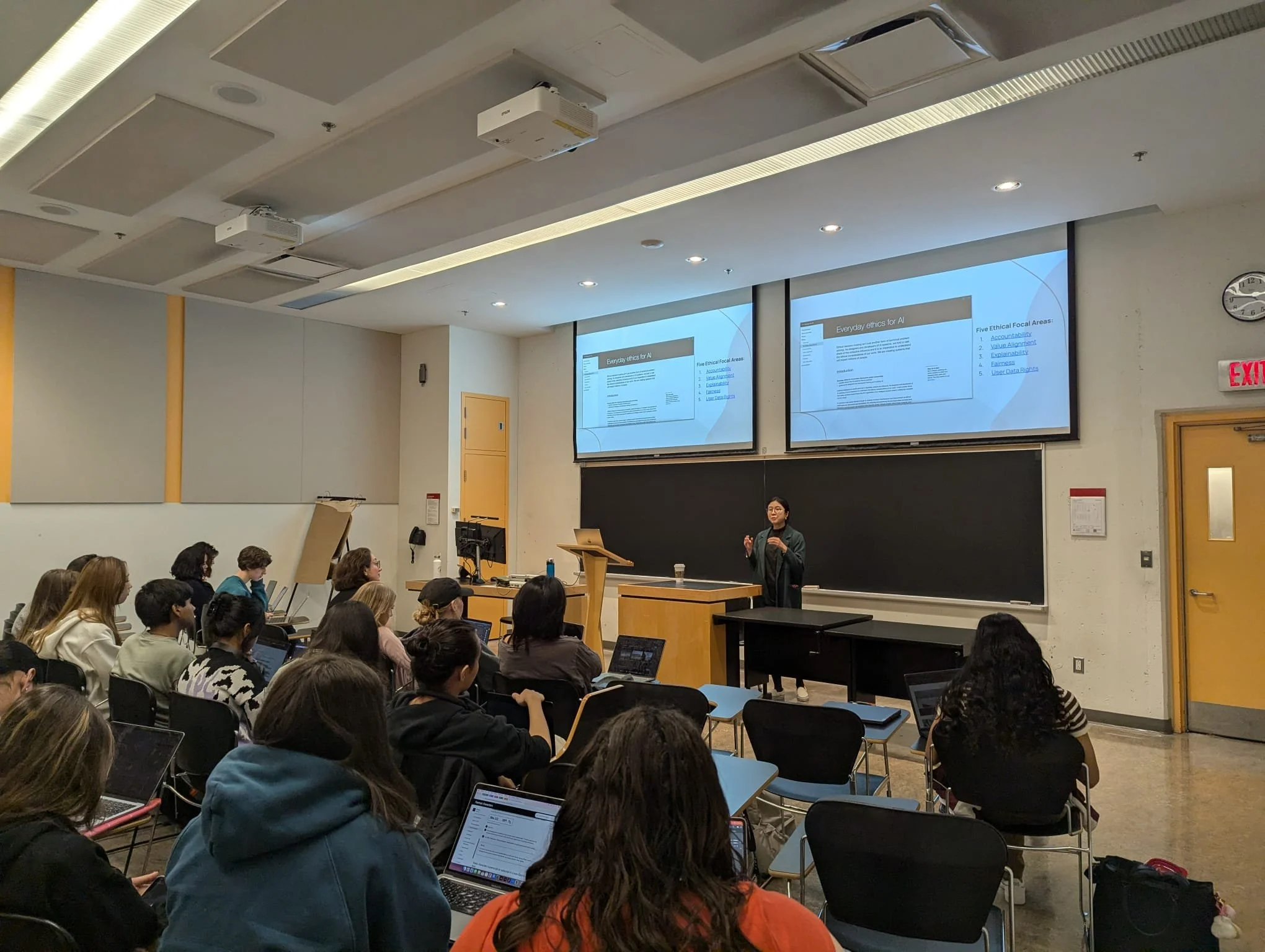 A classroom with students listening to a female instructor giving a presentation at the front. There are two large screens displaying a presentation slide titled 'Everyday ethics for AI.' Multiple students are using laptops and taking notes.
