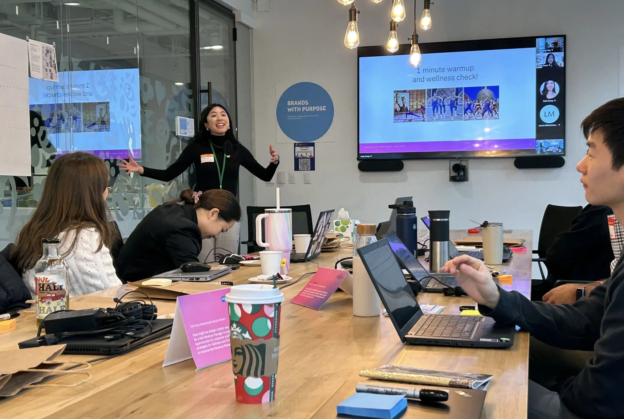 A woman stands at the front of a conference room, giving a presentation to four seated colleagues. The presentation slide on the screen displays a title about wellness checks and images of people exercising. The room has a glass wall, lighting fixtures, and various items like laptops, coffee cups, and notebooks on the table.