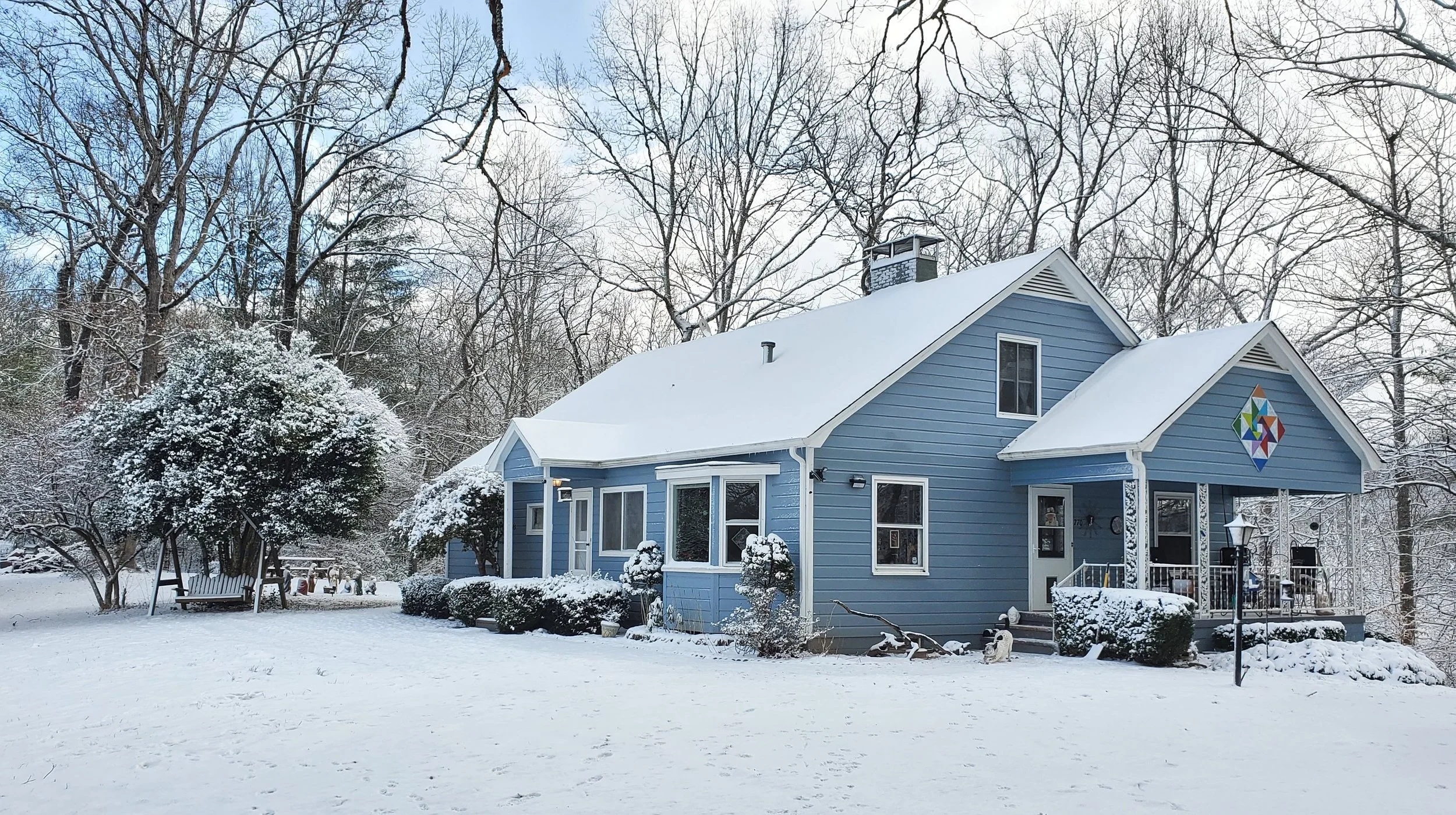A blue house with white trim surrounded by snow-covered trees and bushes in winter, featuring a colorful geometric stained glass window on the front gable, with a snow-covered yard and a swing on the left.