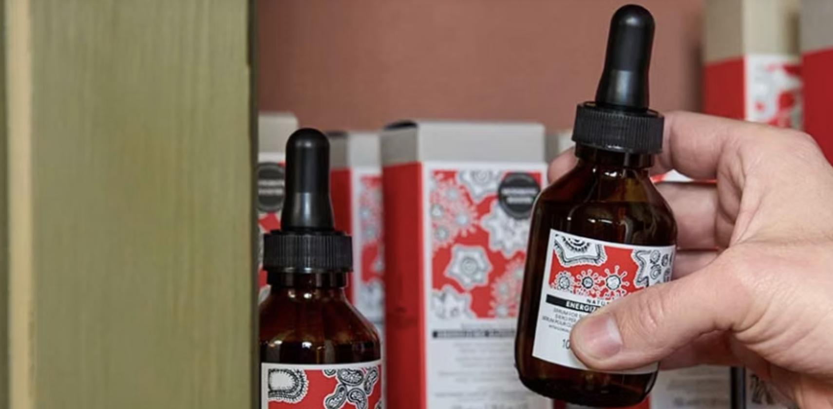 A hand holding a brown glass dropper bottle labeled 'ENERGIZE' next to similar bottles on a shelf.