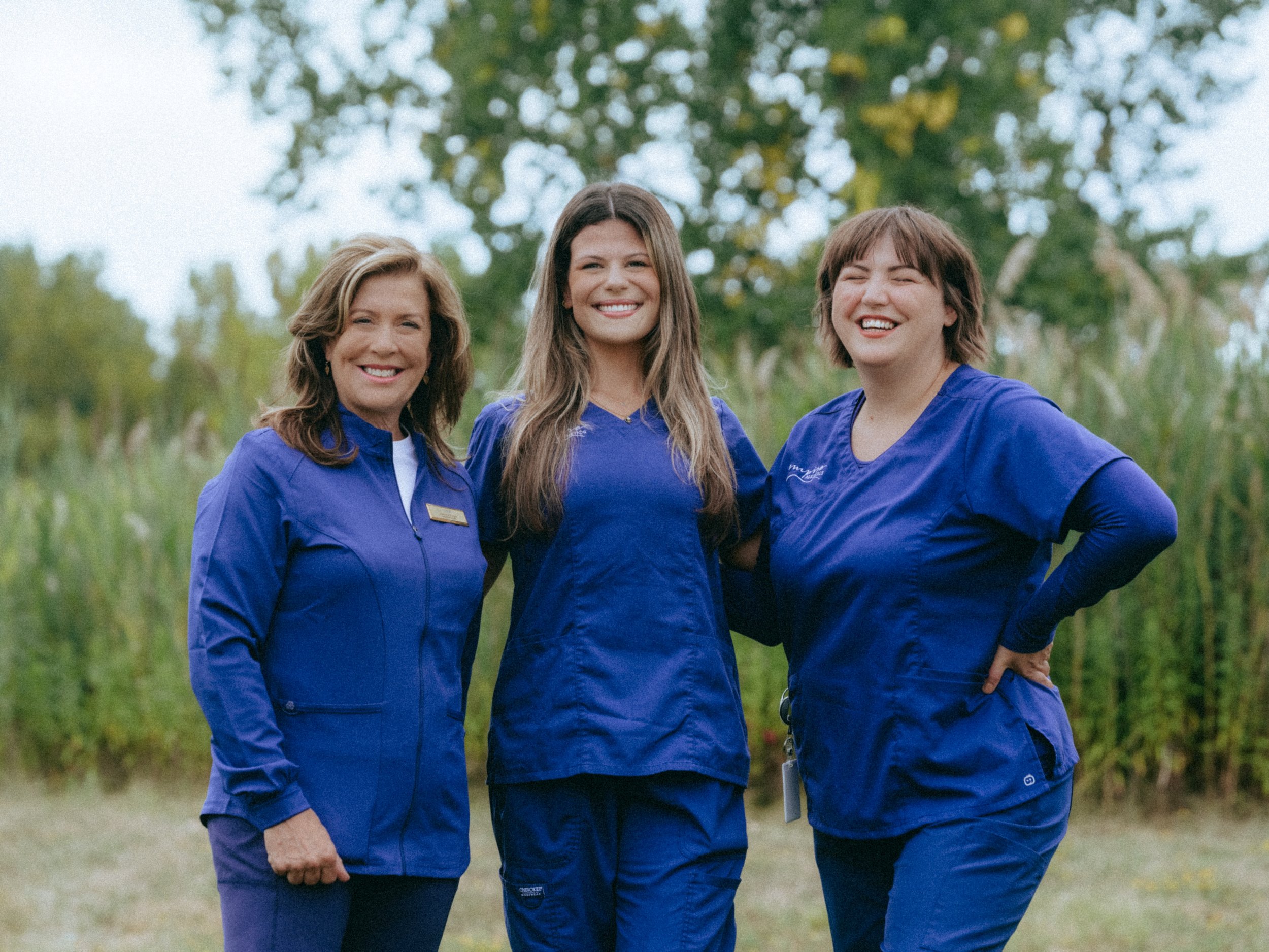 Three women in blue scrubs smiling outdoors with trees in the background.