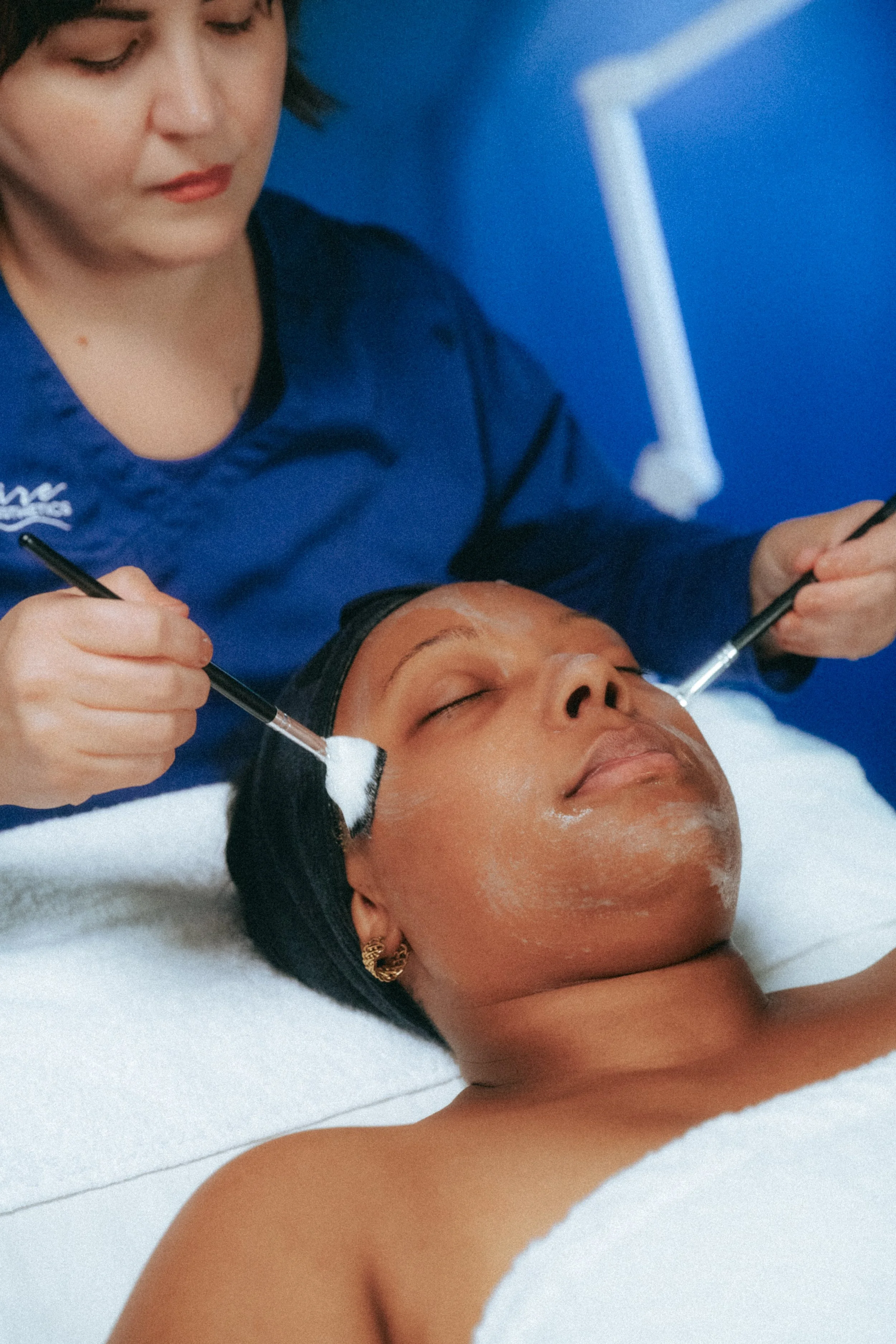 A woman lying on a spa bed receiving a facial treatment, with her eyes closed, while a professional applies skincare products with brushes.
