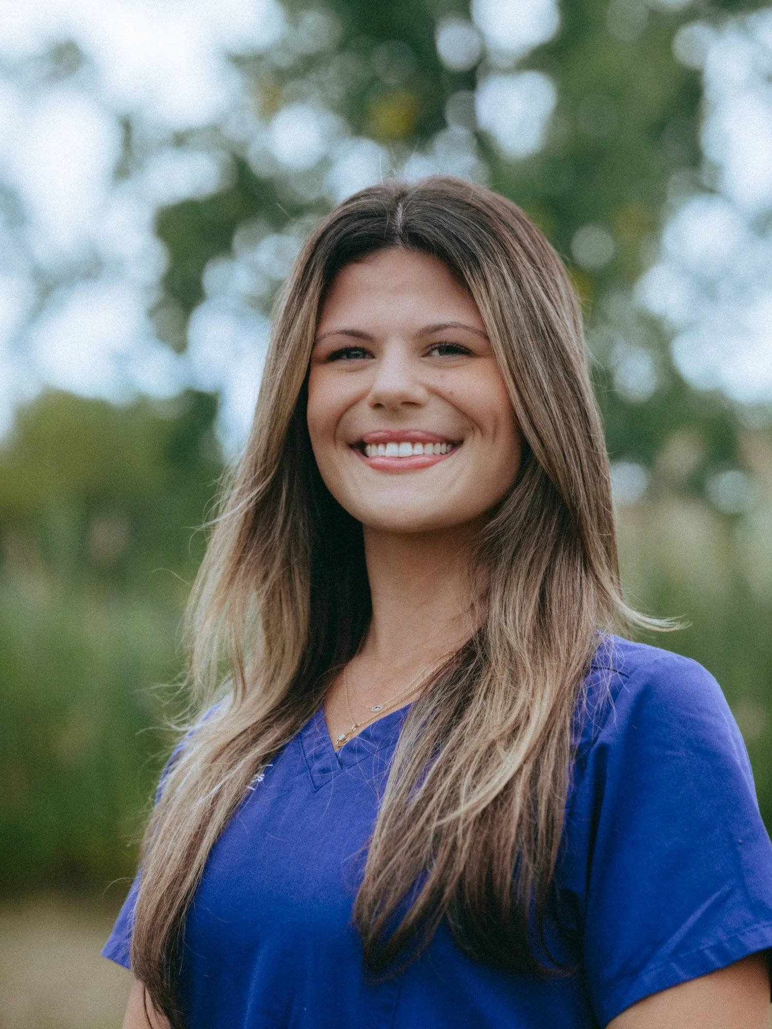 A woman with long brown hair wearing blue scrubs, smiling outdoors with trees in the background.