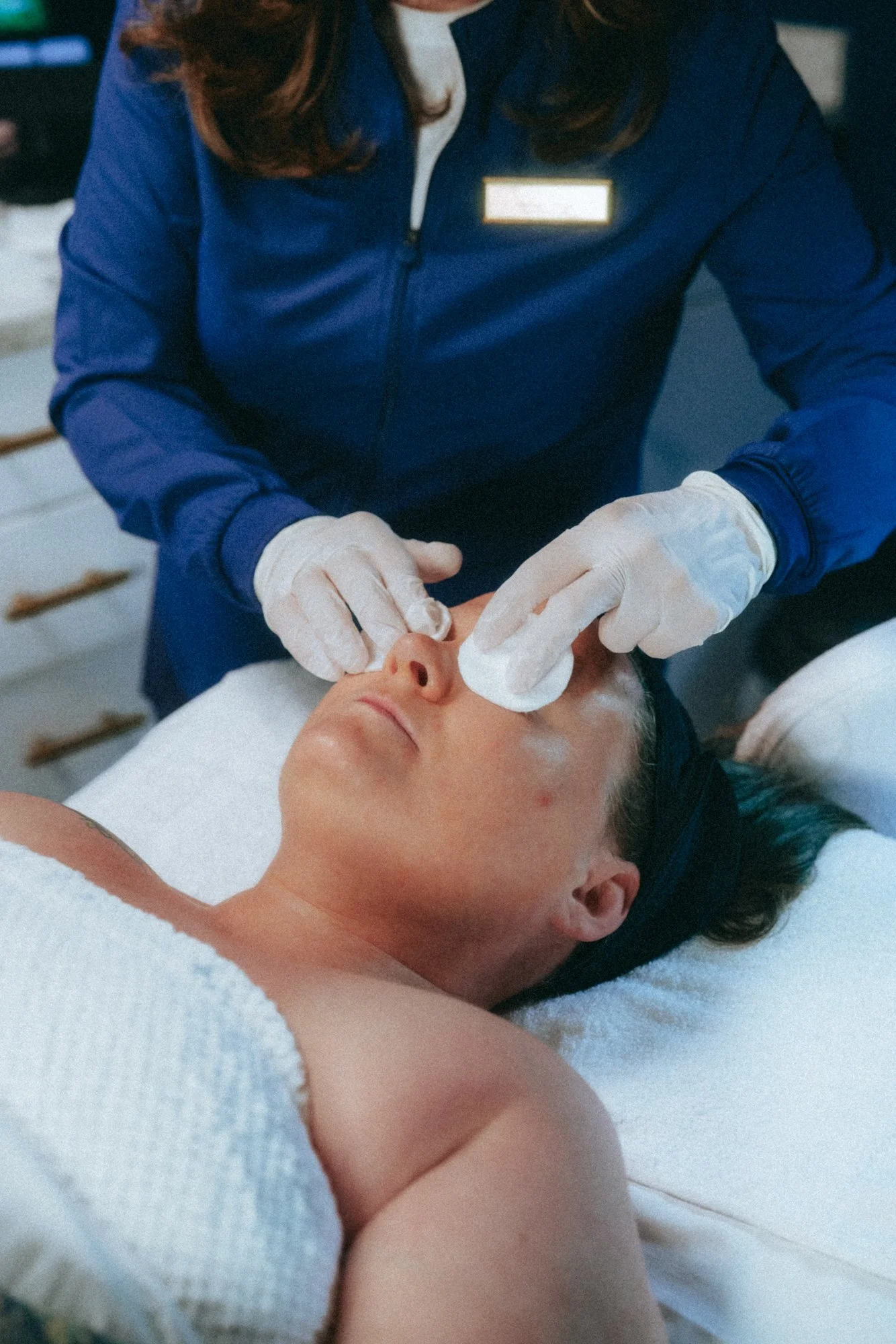 A woman receives a facial treatment at a spa or clinic, lying on a white towel with her eyes closed, as a professional in a blue uniform and white gloves gently cleans her face with cotton pads.
