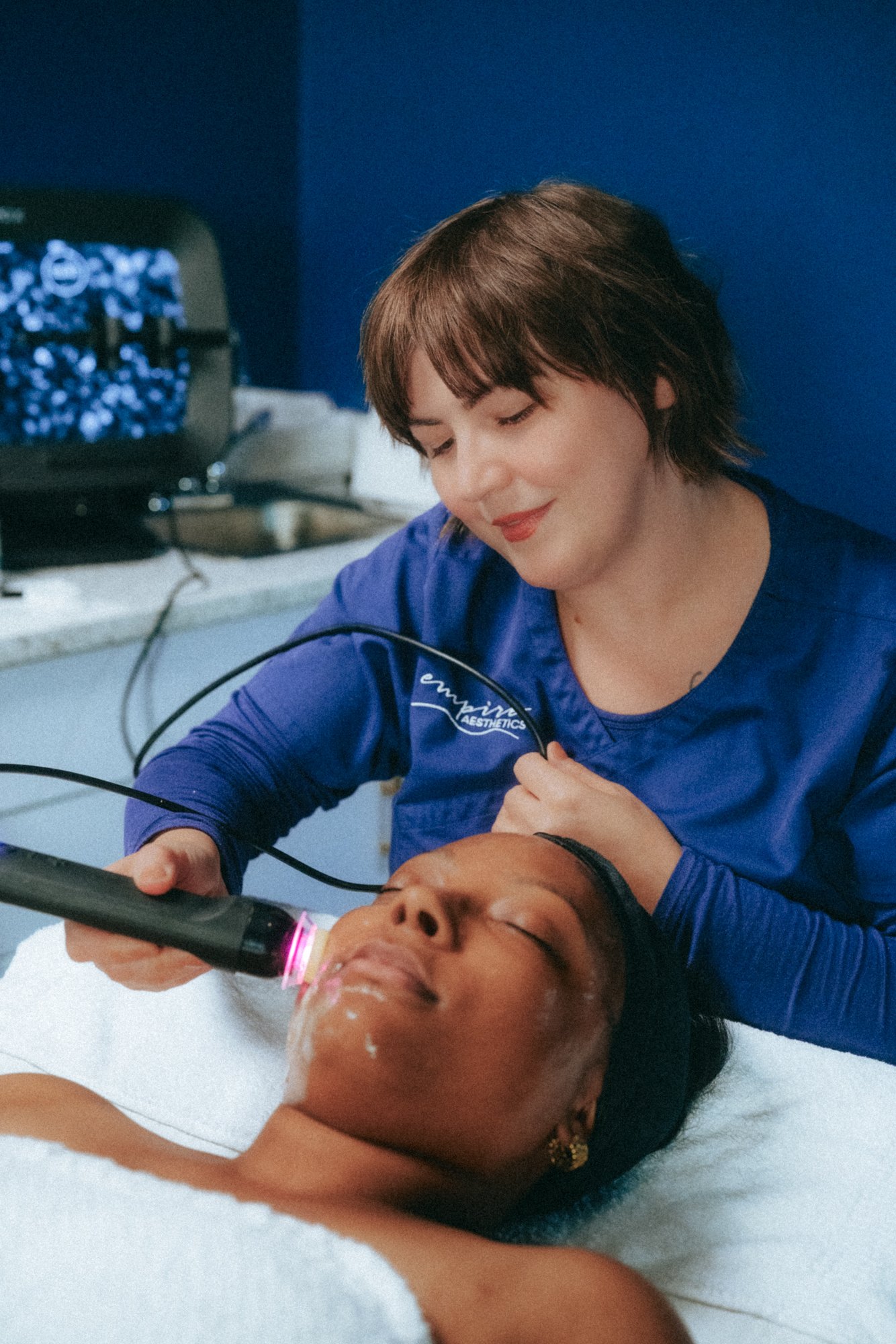 A woman providing a facial treatment with a heated laser device to a woman lying on a treatment bed in a spa or clinic setting.
