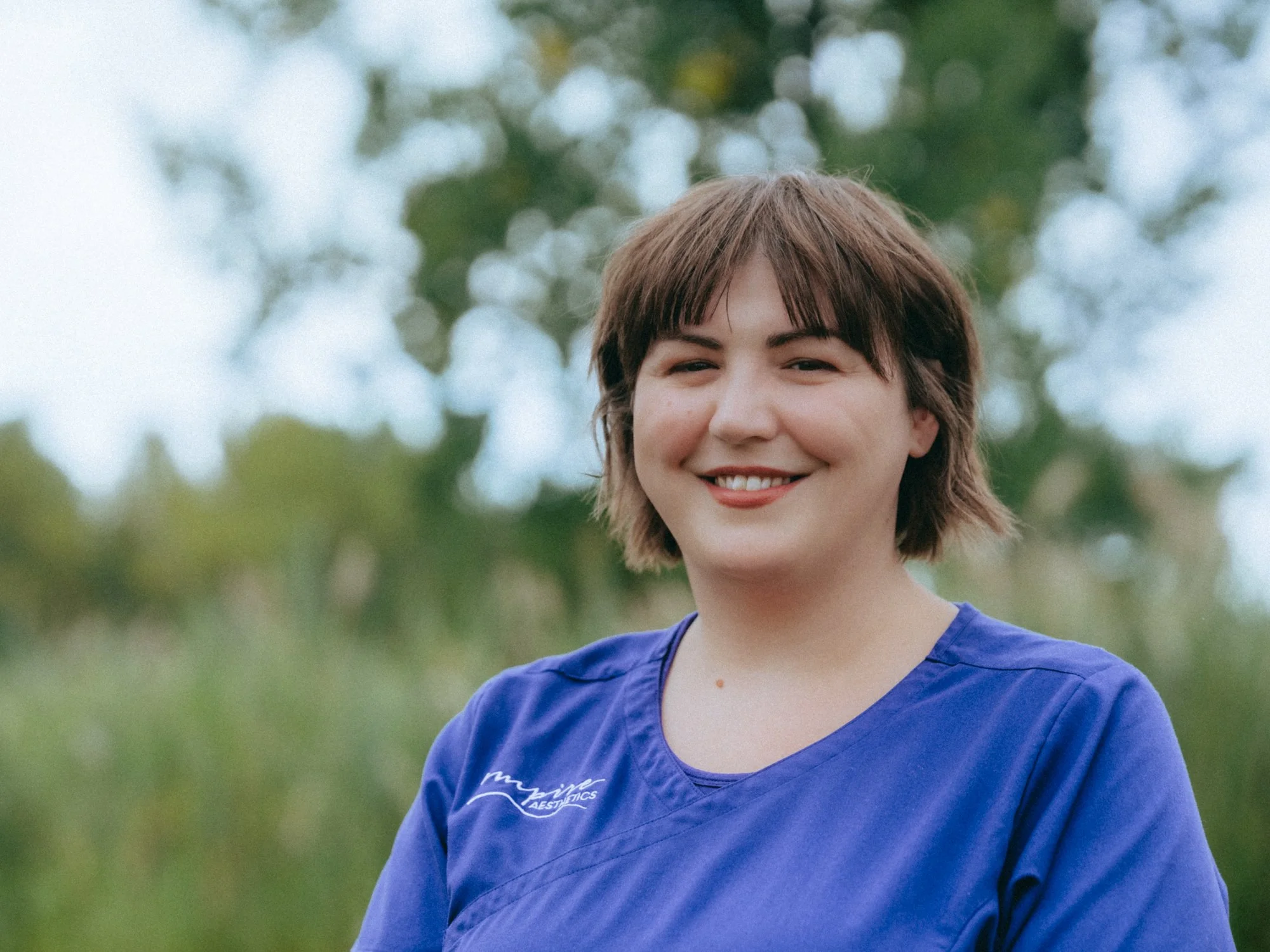 Smiling woman with short brown hair wearing a blue shirt outdoors, blurred green trees in background.