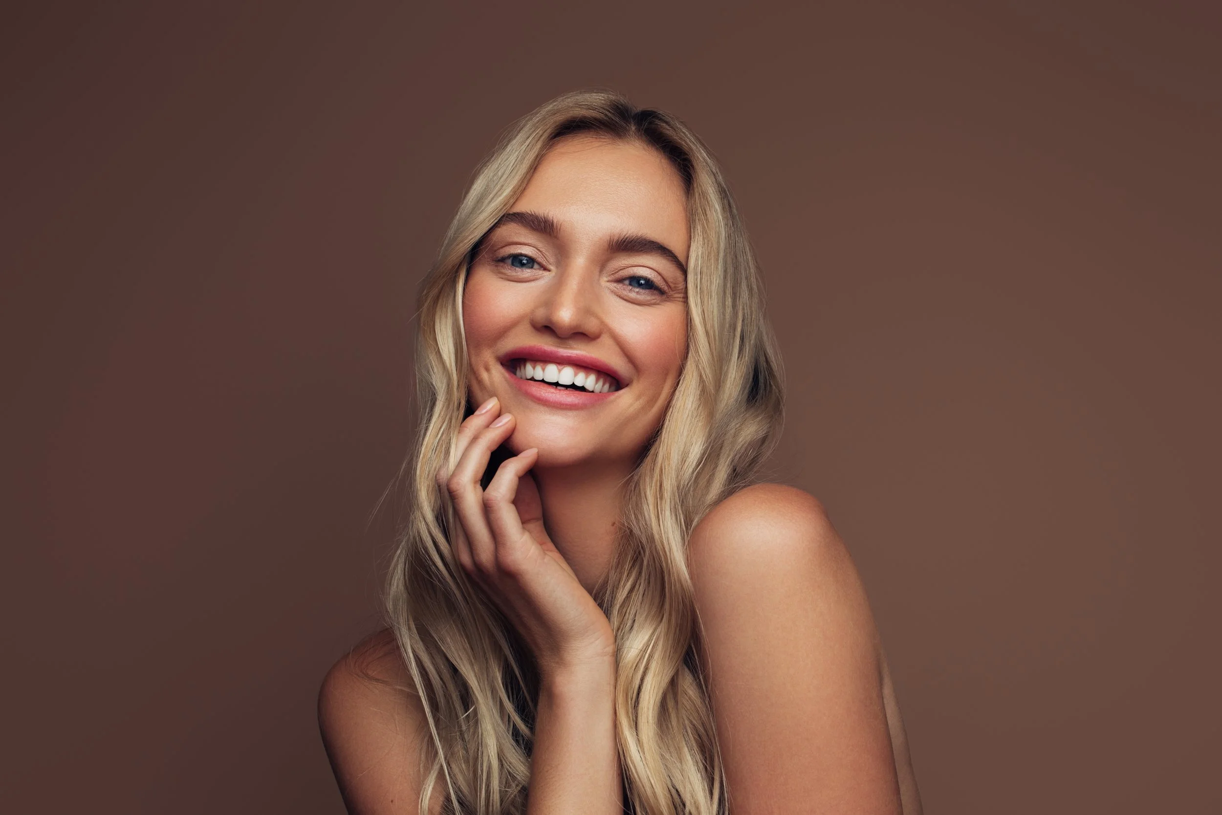 Close-up portrait of a smiling young woman with long blonde wavy hair and blue eyes, touching her face with her right hand, against a plain brown background.