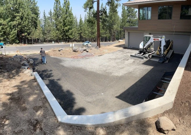 A residential driveway under construction with gravel and a small skid-steer loader parked near the garage, a worker spreading gravel, and a background of trees and parked cars.