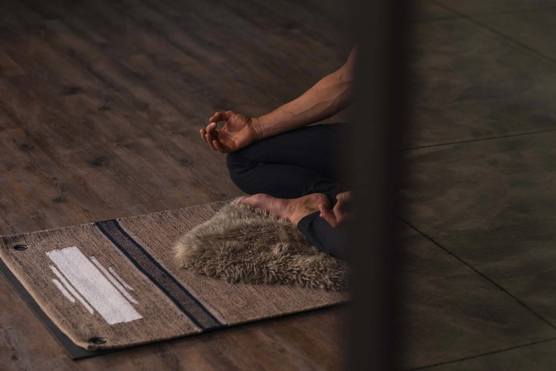 Person practicing yoga in a seated meditation pose on a textured rug with a furry mat, on a wooden floor.