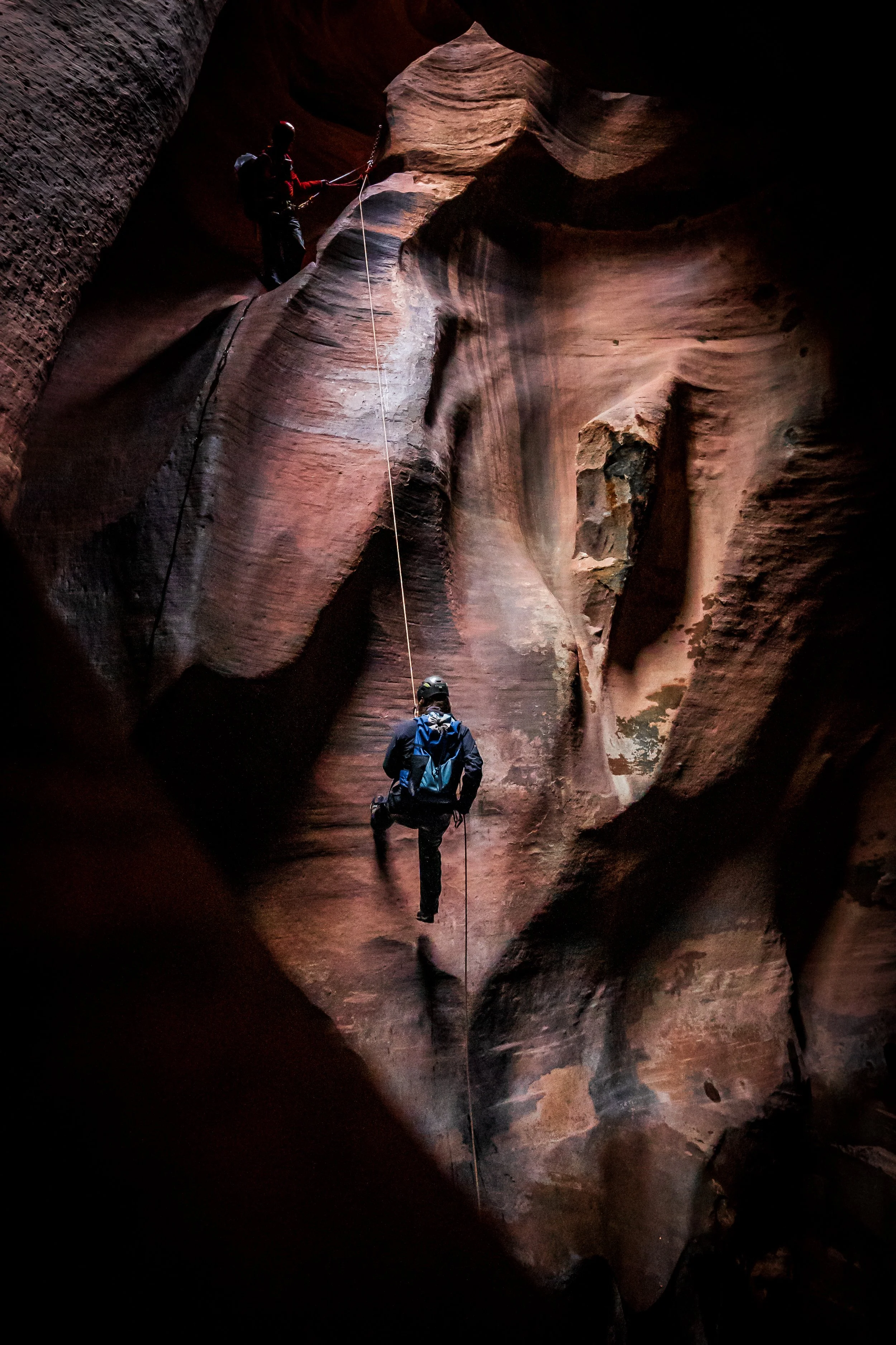Two climbers rappel down a rope inside a narrow red sandstone slot canyon. One stands on a ledge managing the rope while another descends below, illuminated by a beam of light highlighting the canyon’s smooth, sculpted walls.