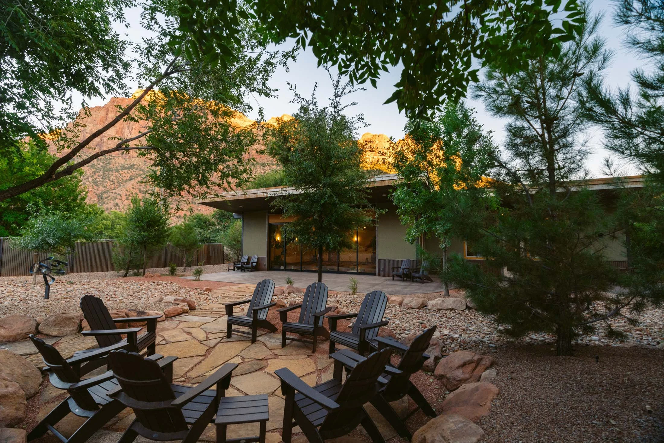 Outdoor patio with black Adirondack chairs arranged in a circle, surrounded by rocks and trees, with a house and mountains in the background.