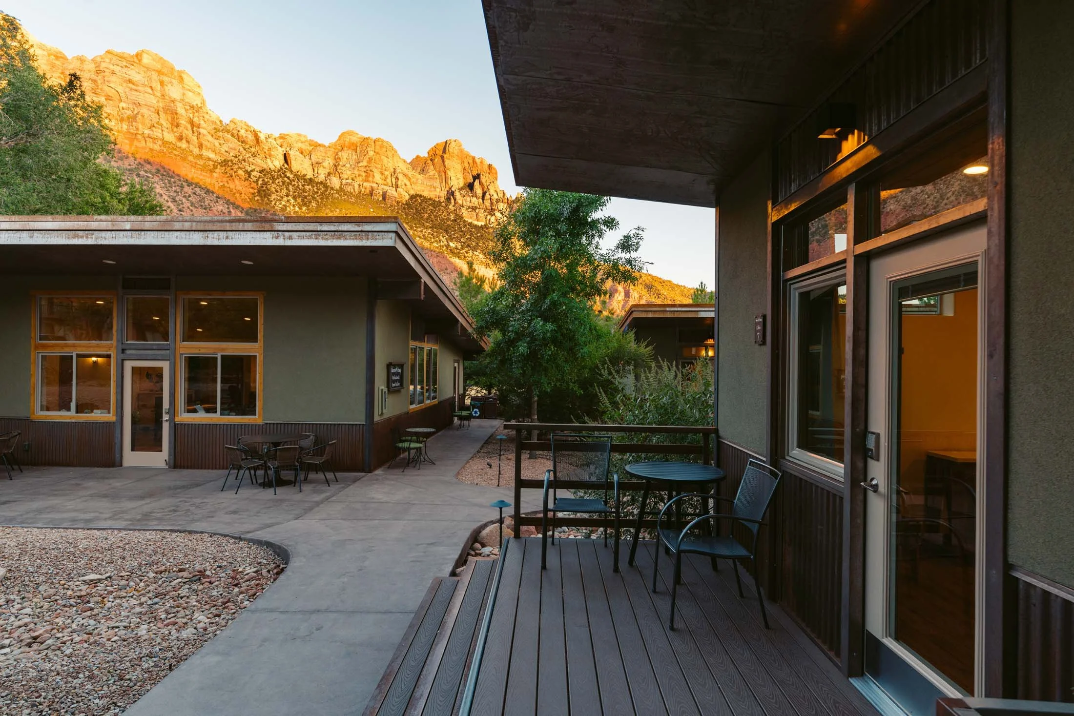 A patio area with outdoor tables and chairs outside of a building. In the background, there are rugged mountains illuminated by the setting sun.