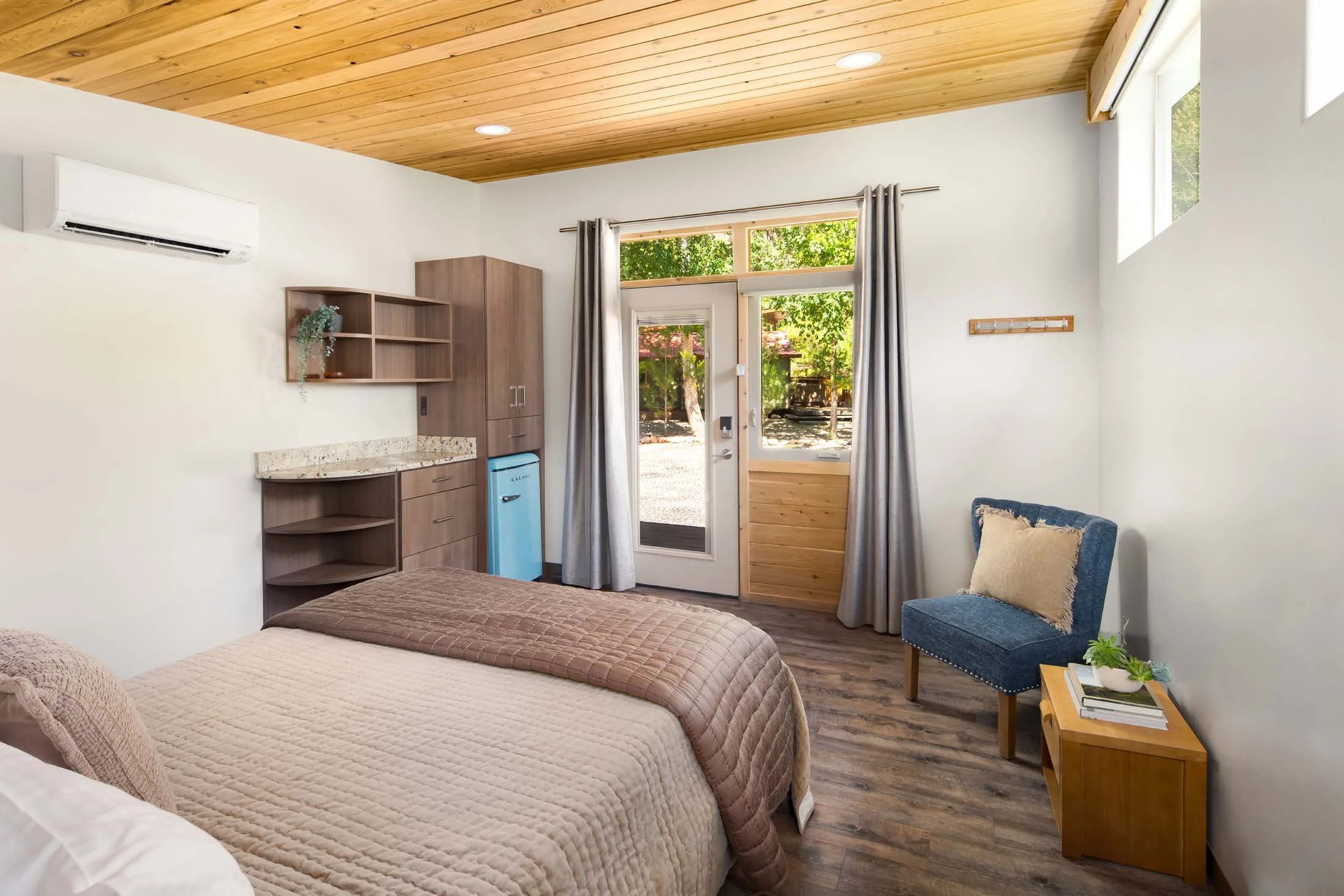 Rooms in Zion National Park - A cozy bedroom with a bed, blue chair, small wooden side table with books and a plant, a door leading outside, and a wooden ceiling.