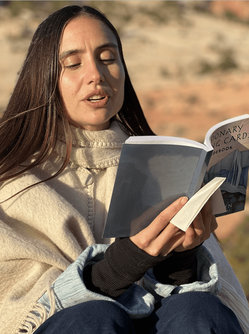 A woman with long brown hair and light skin, wearing a cream-colored sweater, reading a book outdoors during sunset with a blurred natural landscape in the background.