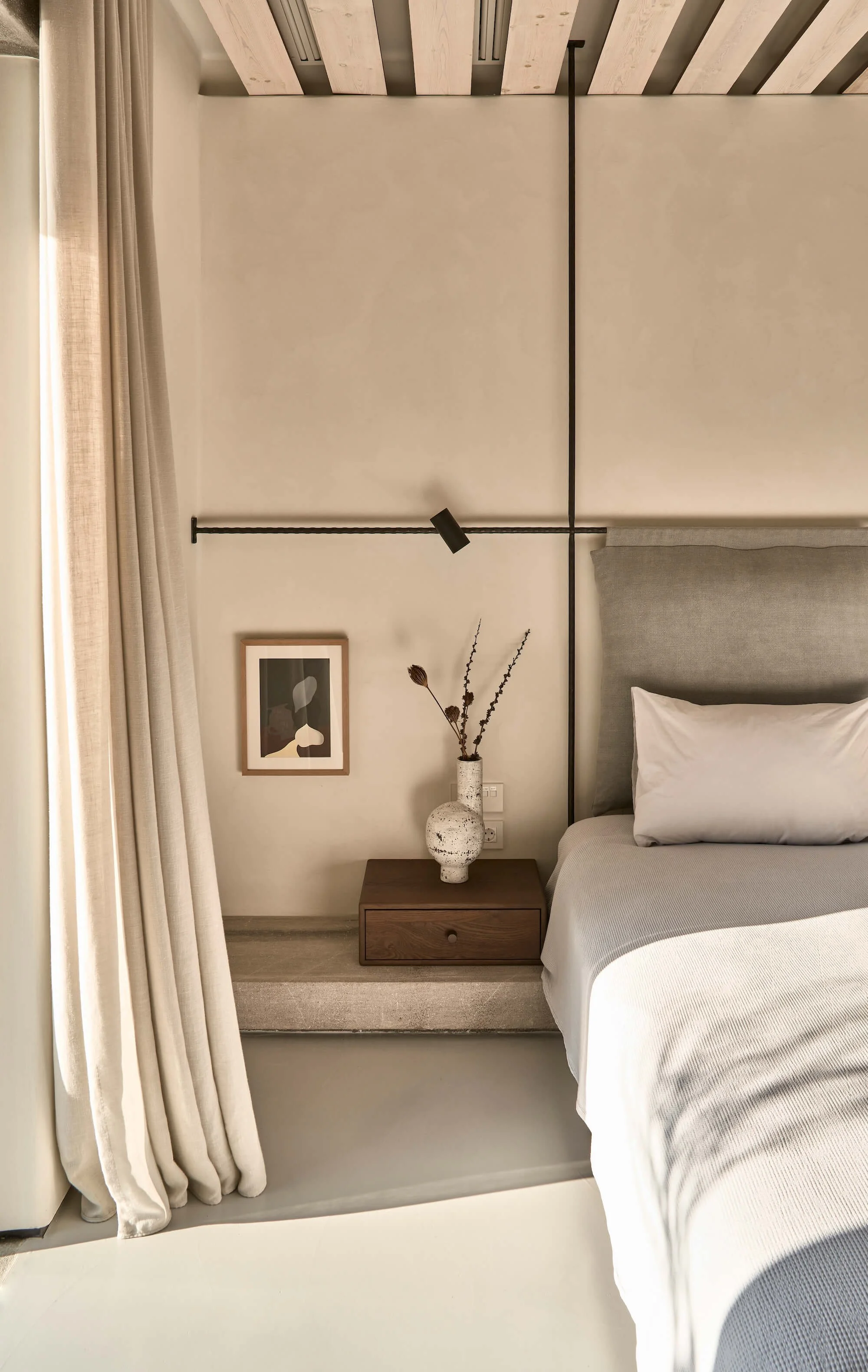 A minimalist bedroom with a bed, a nightstand with a vase and dried flowers, a wall-mounted light, a framed artwork, and beige curtains.