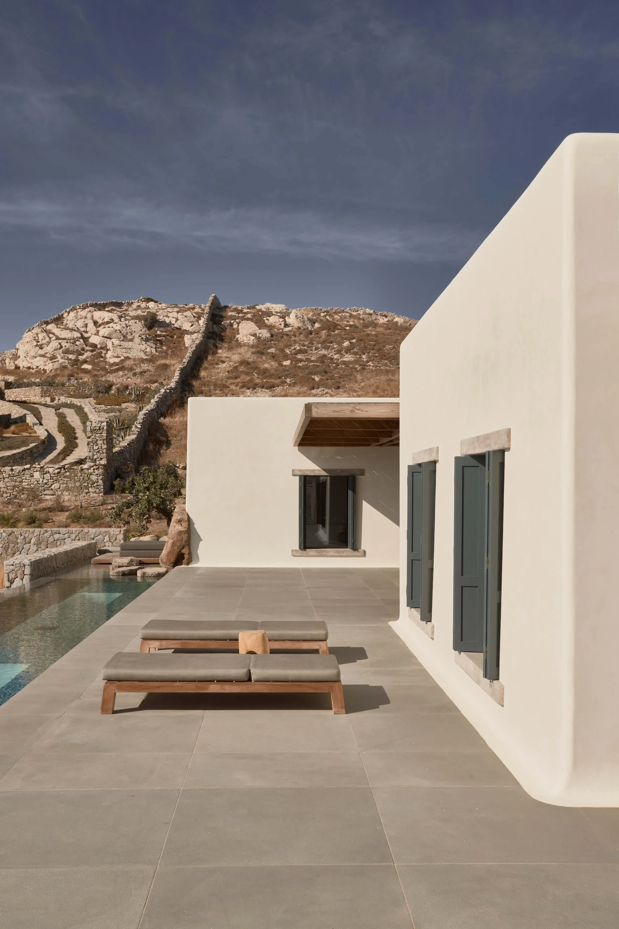 Modern villa with a white curved exterior, blue shutters, wooden outdoor furniture on a tiled patio, a swimming pool, rocky hillside with terraced stone walls, and a clear sky.