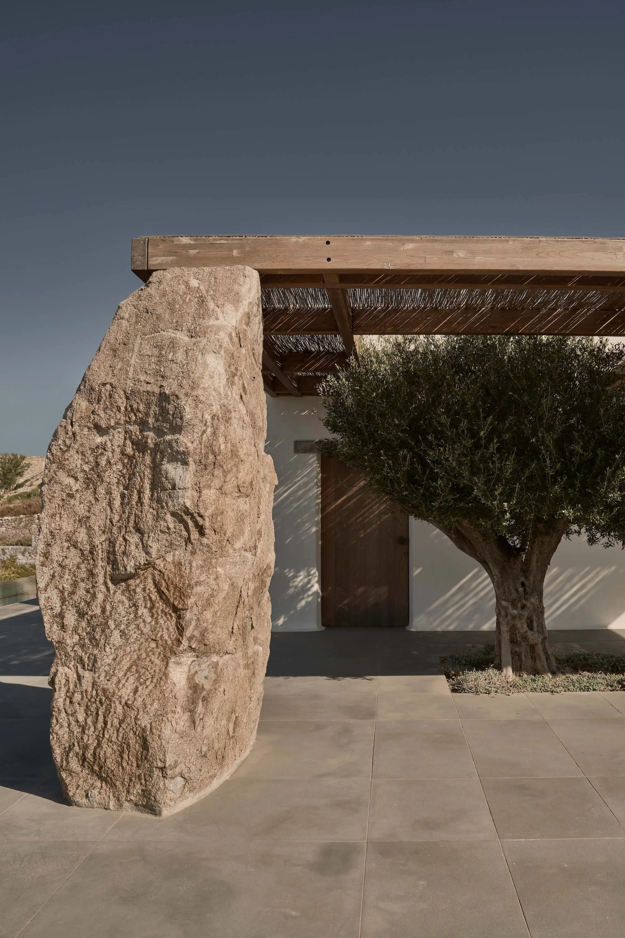 A minimalist modern outdoor scene with a large, rough-textured stone, a tree with dense foliage, and a building with a wooden door and an overhang made of wood and reeds. Clear sky in the background.