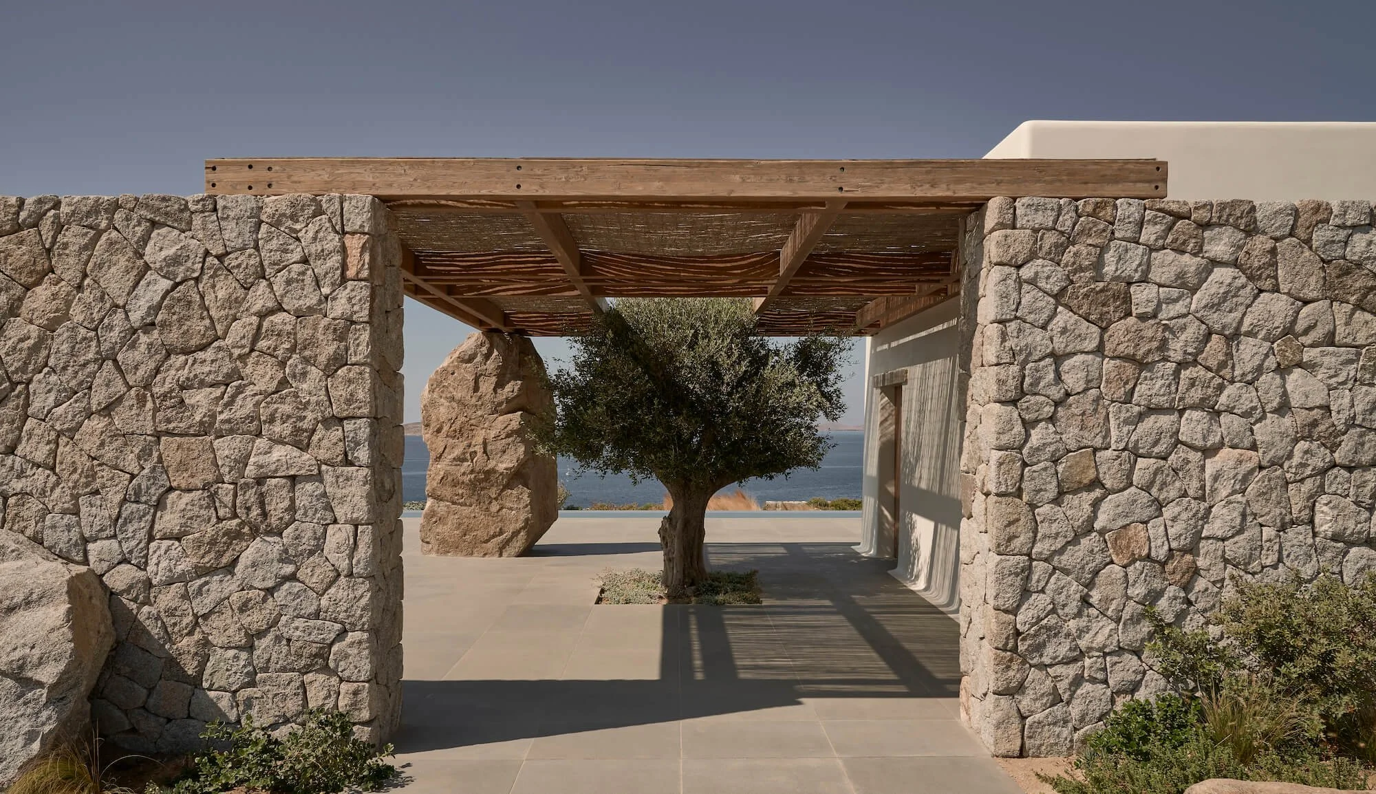 Stone and wood pergola overlooking the ocean with a tree in the center and a large rock in the background.