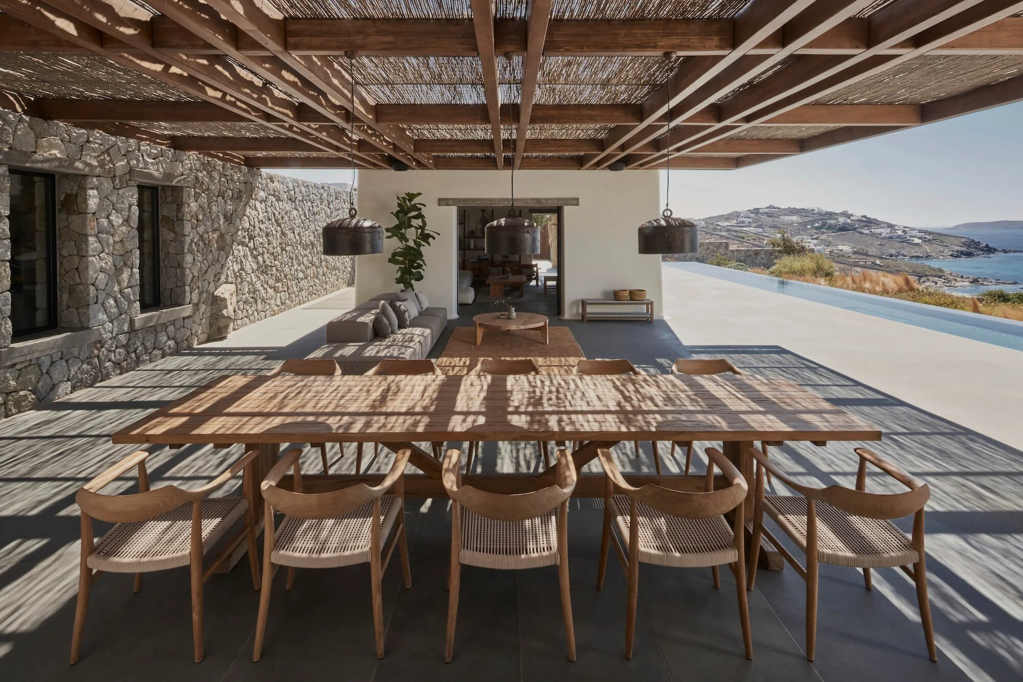 Modern outdoor patio with a large wooden dining table and nine chairs, stone wall, outdoor sofa with pillows, coffee table, and view of a coastline with hills and water in the distance.