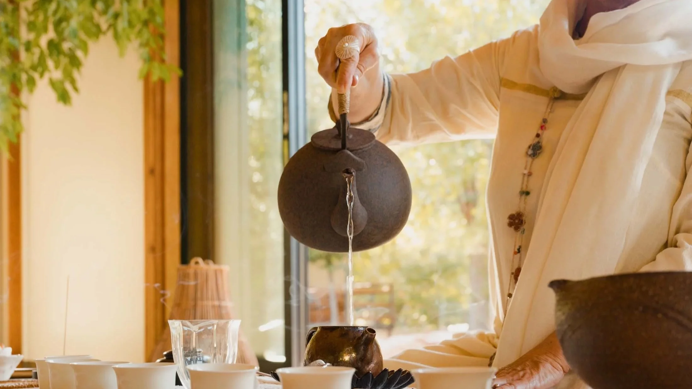 Person pouring tea from a teapot into cups on a table near a window with fall foliage outside.