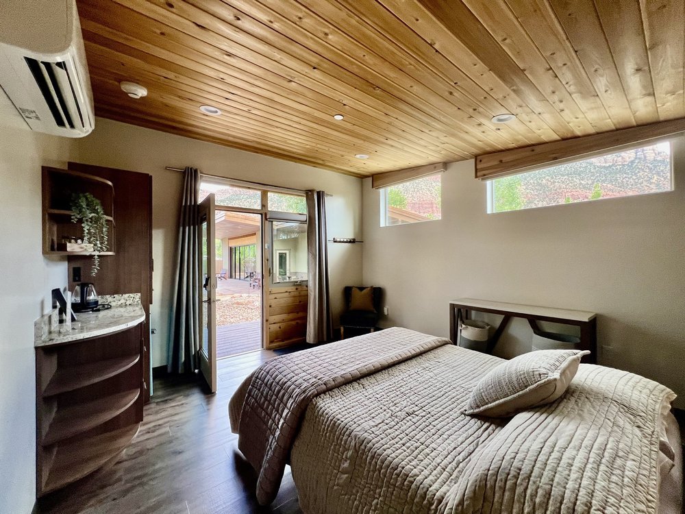A cozy bedroom with a wooden ceiling, a bed with beige bedding, a small desk, a chair, and a door opening to an outdoor deck with trees.
