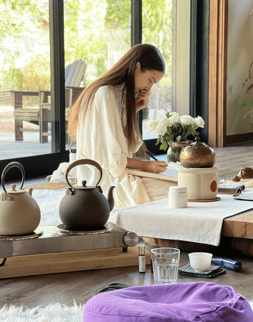 A woman with long brown hair sitting on the floor near a low table, writing in a notebook, with teapots, a flower vase, and various items on the table, near a large glass door showing an outdoor patio and trees.