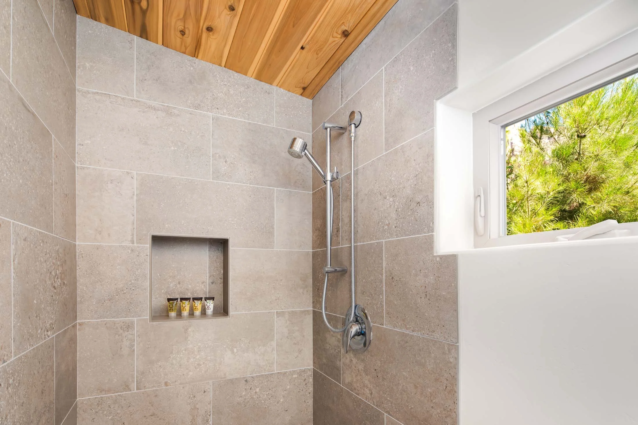 A modern shower with beige tiled walls, a wood ceiling, a built-in shelf with small bottles, a silver adjustable shower head, a window showing greenery outside.