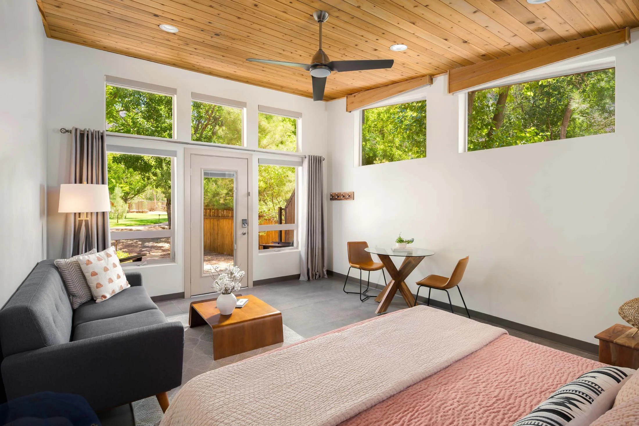 Living room with large windows showing greenery outside, beige walls, a wooden ceiling fan, a gray sofa with pillows, a wooden coffee table, a small dining table with two chairs, and a door leading outside.
