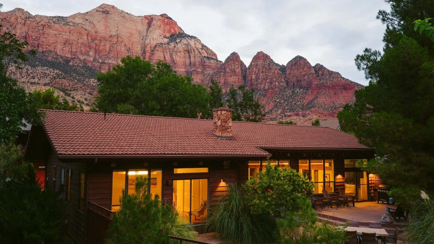 A house with a red tile roof and large glass windows, surrounded by trees, with red mountains in the background during dusk.