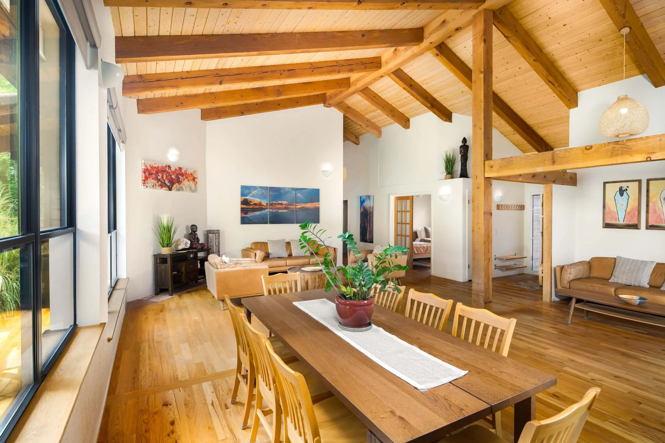 Interior of a living and dining room with wooden beams and flooring, a large dining table with a potted plant, beige sofas, artwork on the walls, and large windows with natural light.