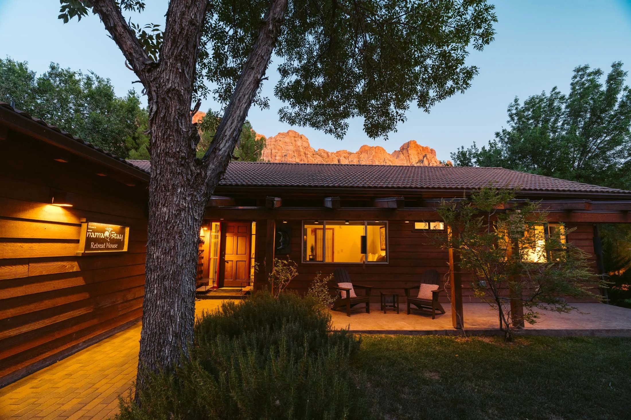 A cozy retreat house at dusk with warm lighting, surrounded by trees and plants, with mountain cliffs in the background.