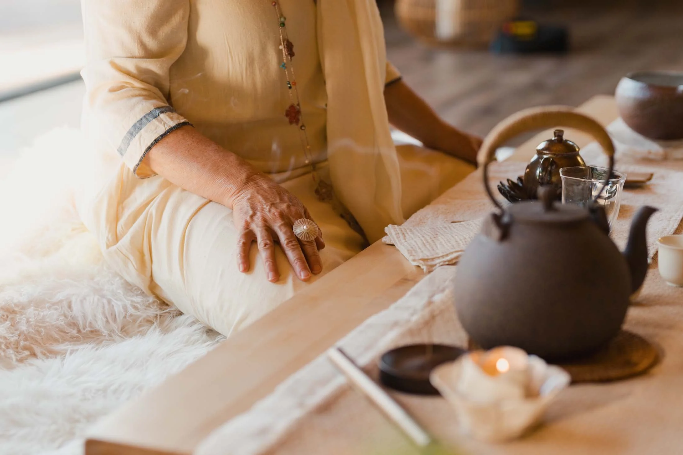 A person in cream clothing sits beside a low tea ceremony table with teapots, cups, soft linens, and candlelight.