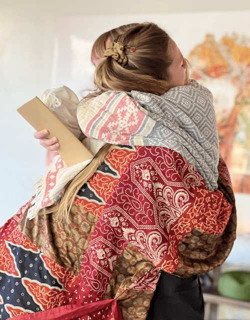 Two women hugging each other warmly, one dressed in colorful, patterned clothing and the other in gray, with a gift box in hand, in an indoor setting.