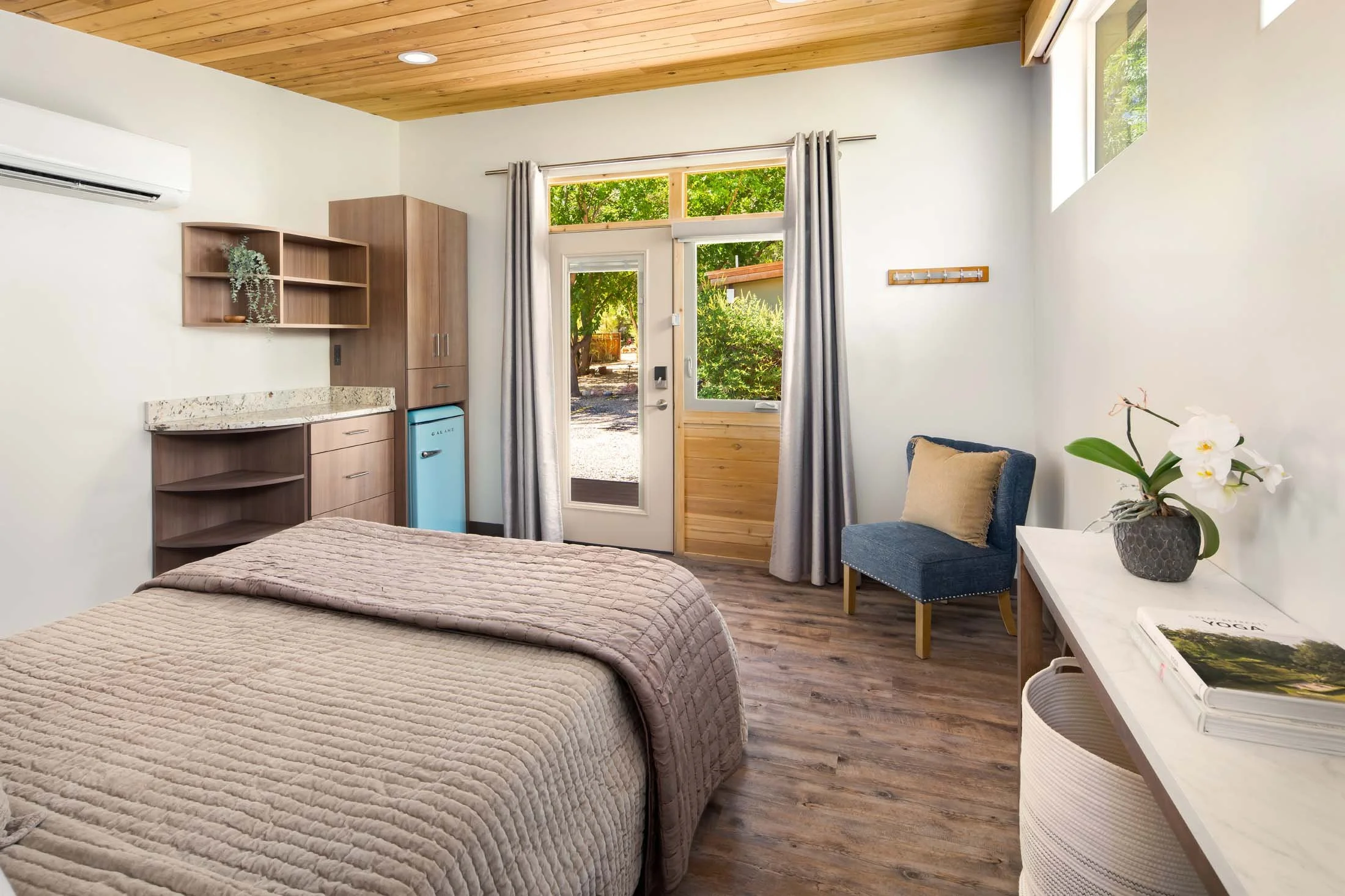 Rooms in Zion National Park - A cozy bedroom with a wooden ceiling, beige quilted bed, blue chair, marble-topped desk with flower vase, and a door with a window leading outside.