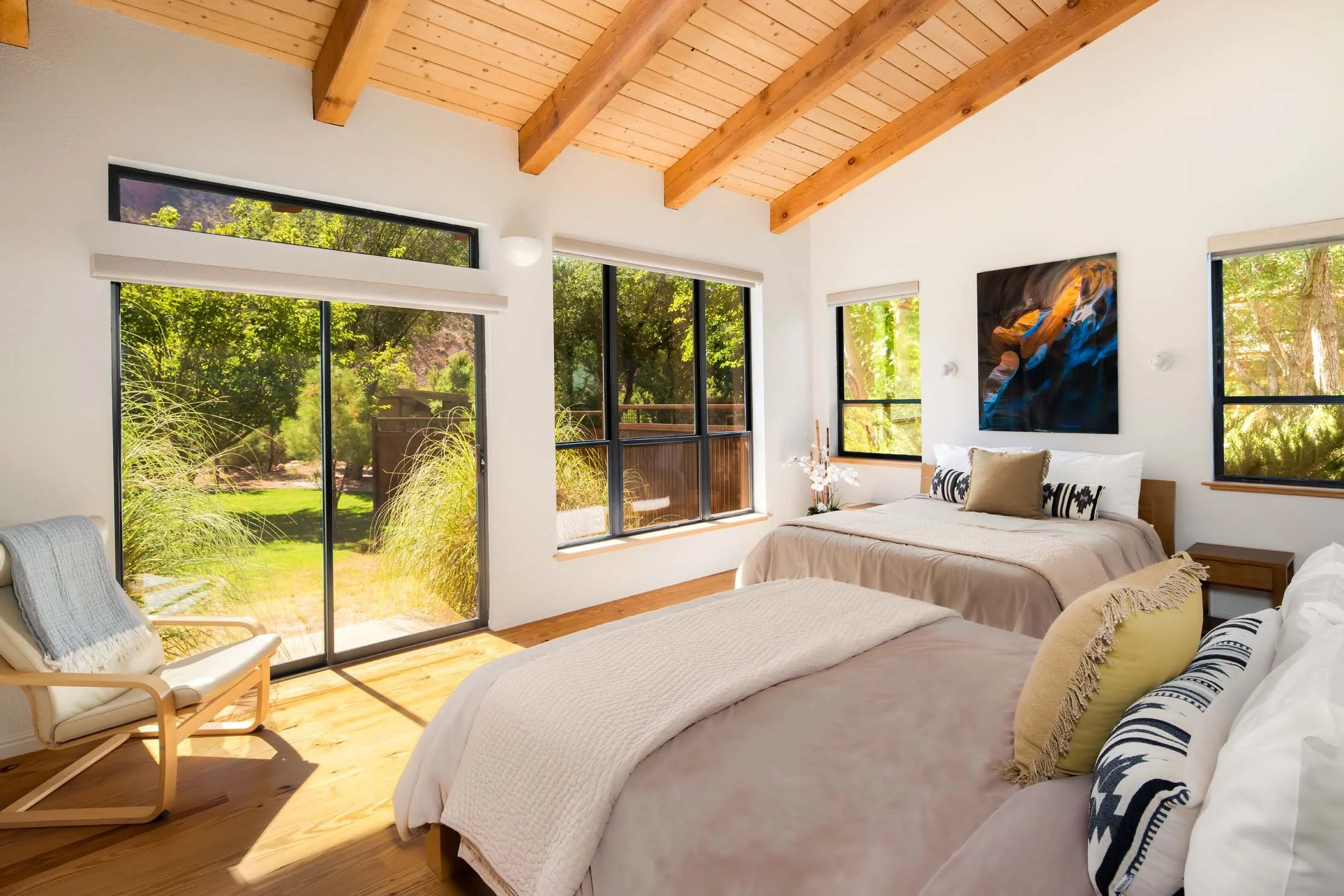 Bright bedroom with two beds, large windows showing greenery, wooden ceiling with exposed beams, and modern decor.