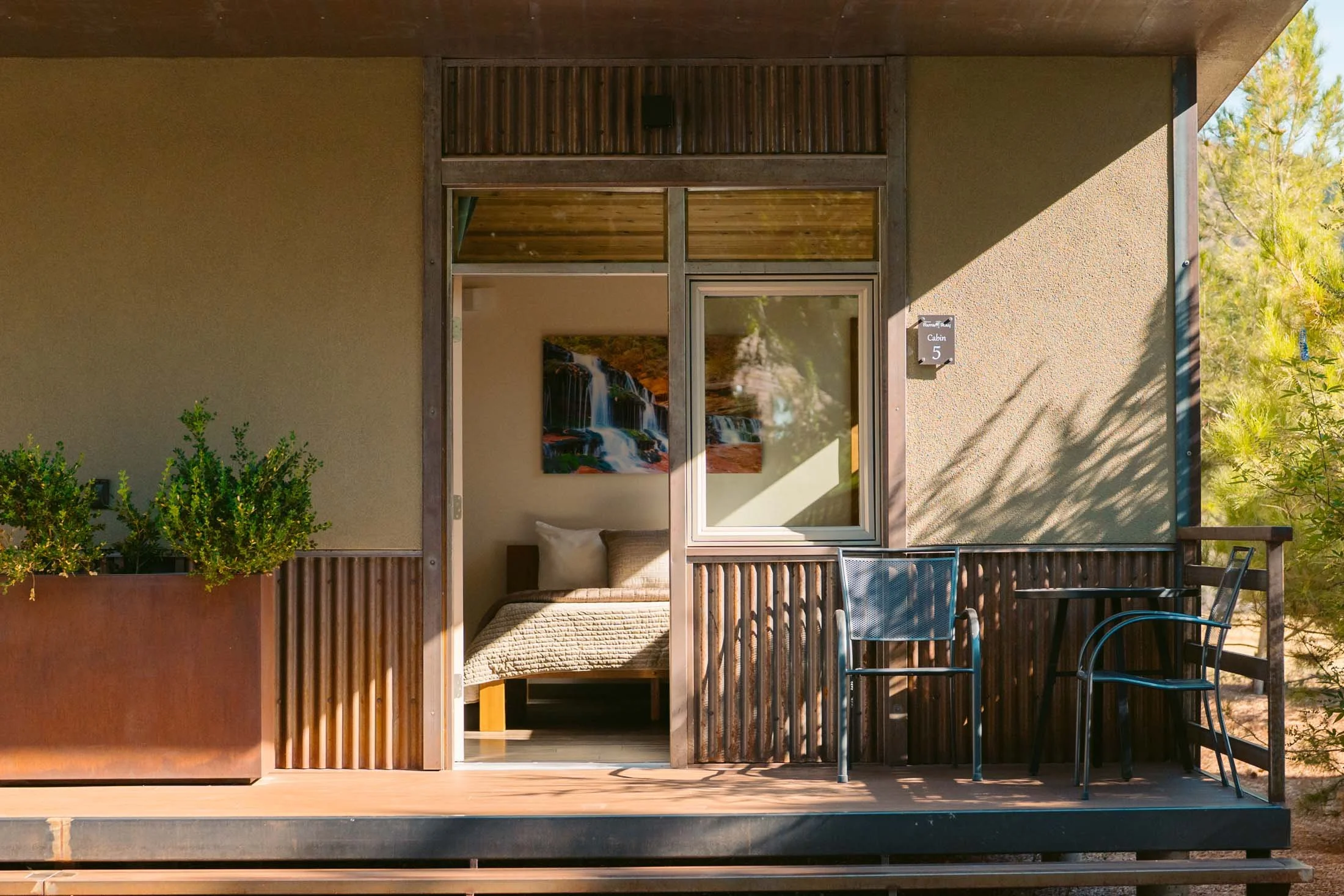 Exterior view of a cabin with a small porch. The porch has two black chairs and a small table, with a potted plant on the left. The door is open, revealing a bed inside with a painting of waterfalls on the wall. There is a sign that reads 'Cabin 5' o