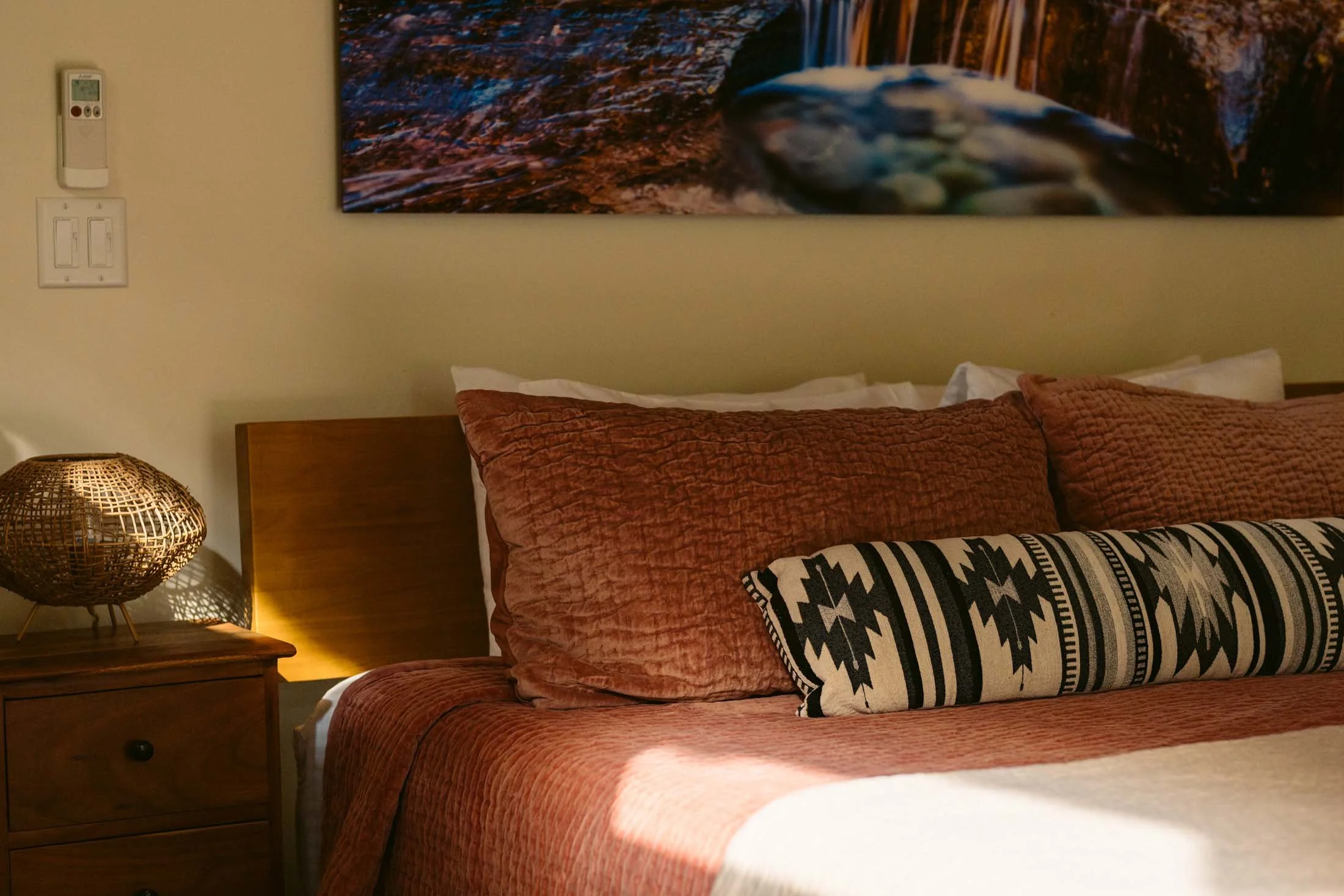 A cozy bedroom corner featuring a bed with rust-colored bedding and pillows, a patterned bolster pillow, a wooden nightstand with a decorative wicker lamp, and wall art depicting a scenic landscape with water and distant trees.