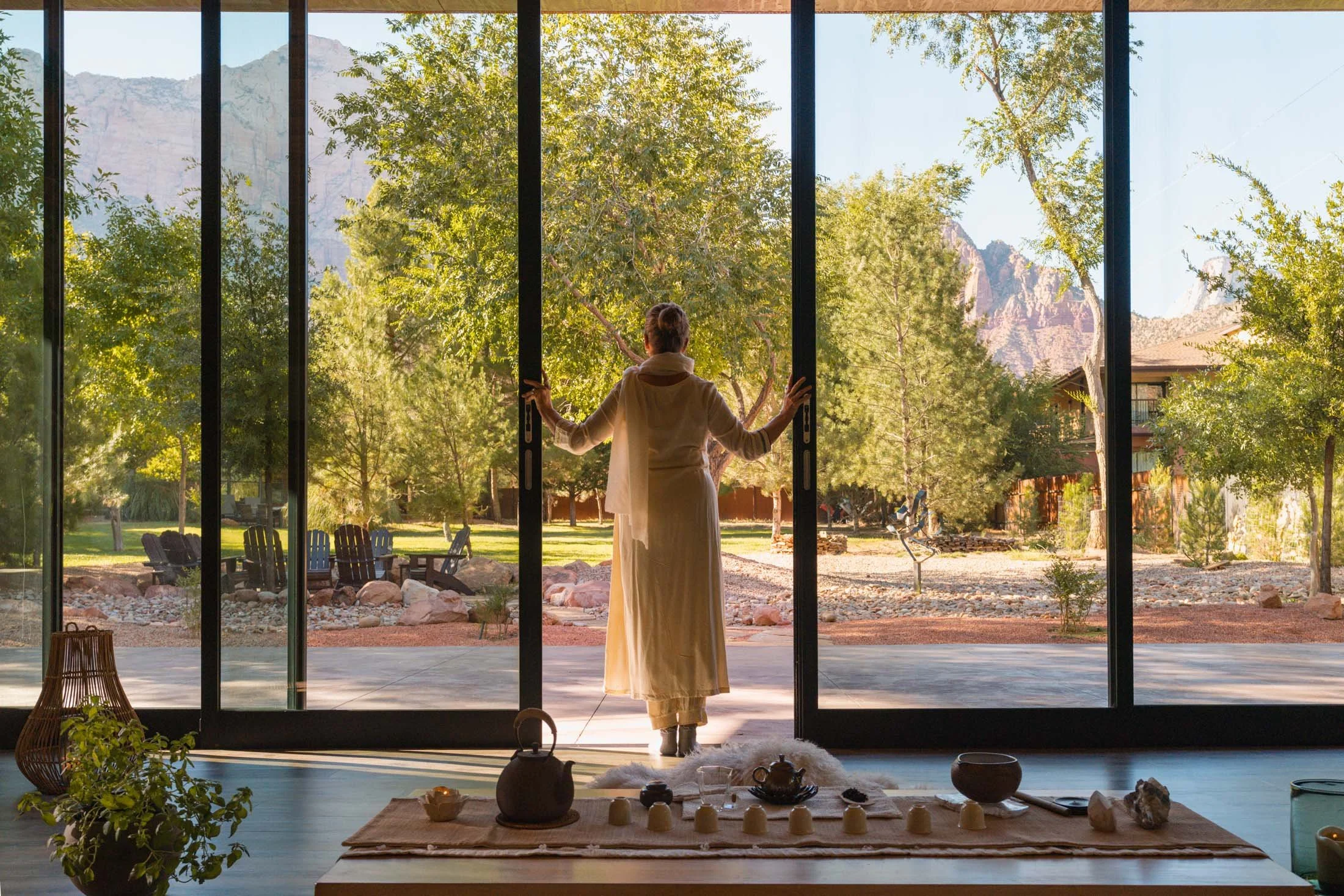 A woman opening glass sliding doors to a sunny outdoor patio with trees and mountains in the background. Inside, there is a tea set and various cups on a beige rug. Property of Nama-Stay, offering vacation rentals in Springdale, Utah.