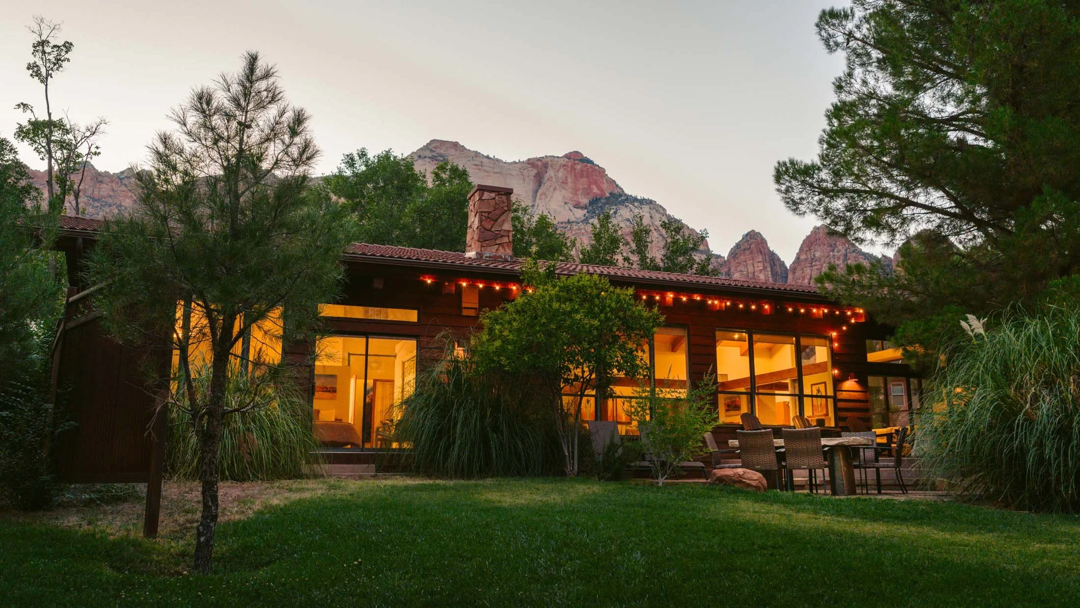 A house with large glass windows, surrounded by trees and greenery, with a mountain range in the background, and string lights hanging from the roof at dusk.