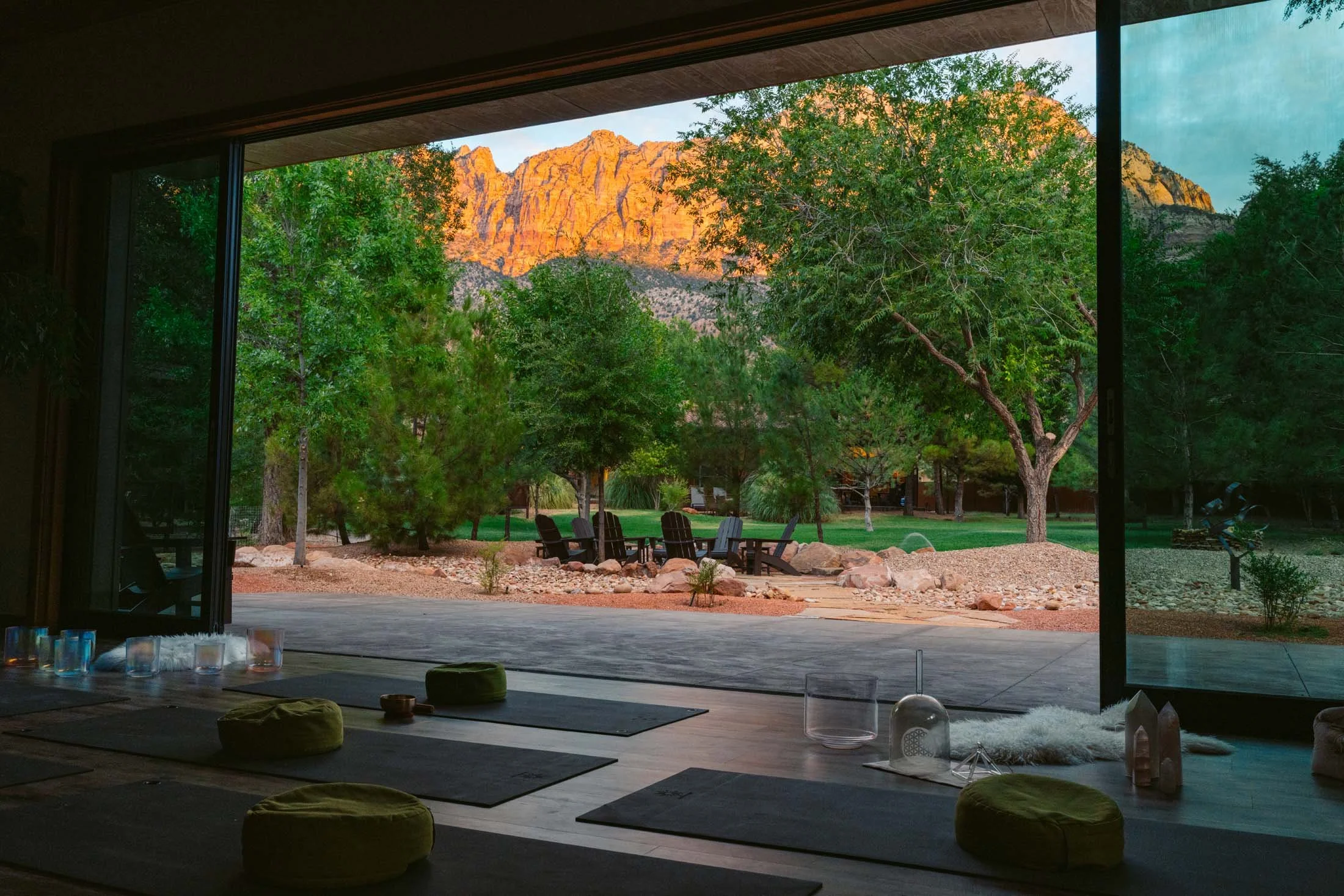 Interior view of a yoga or meditation room with mats and cushions, overlooking a lush outdoor garden with trees and mountain scenery in the background.