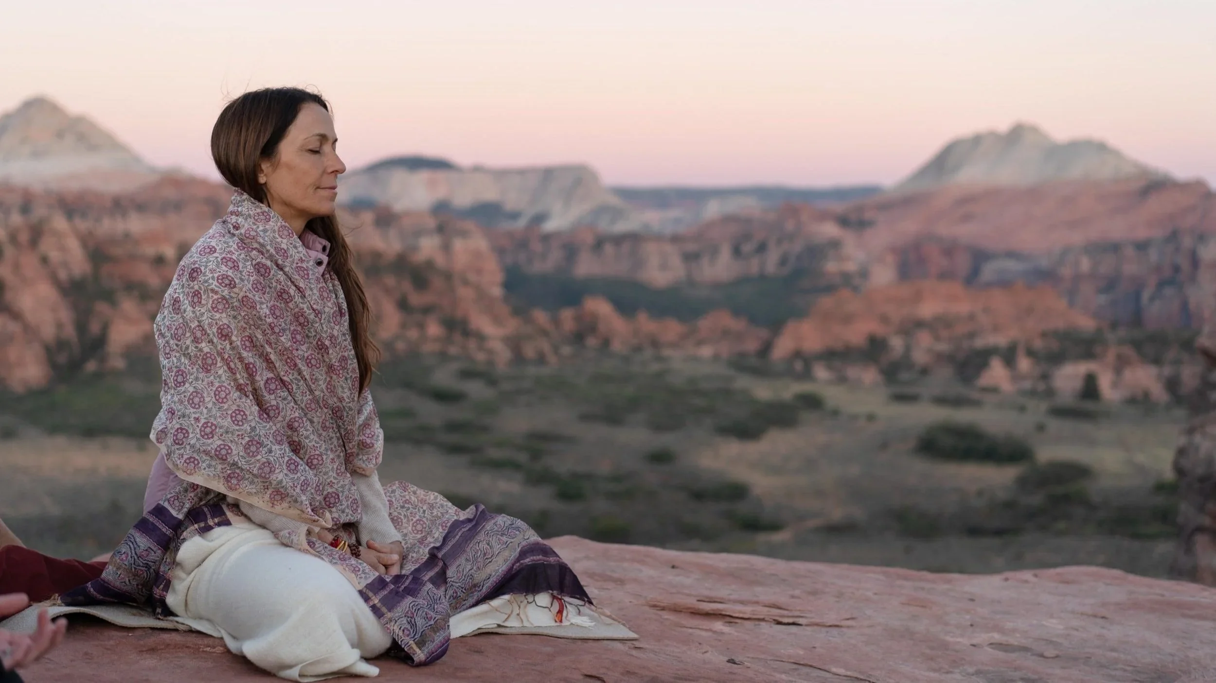 A woman in patterned clothing sitting peacefully in a meditative pose on a rock at sunset, with a desert canyon landscape in the background.