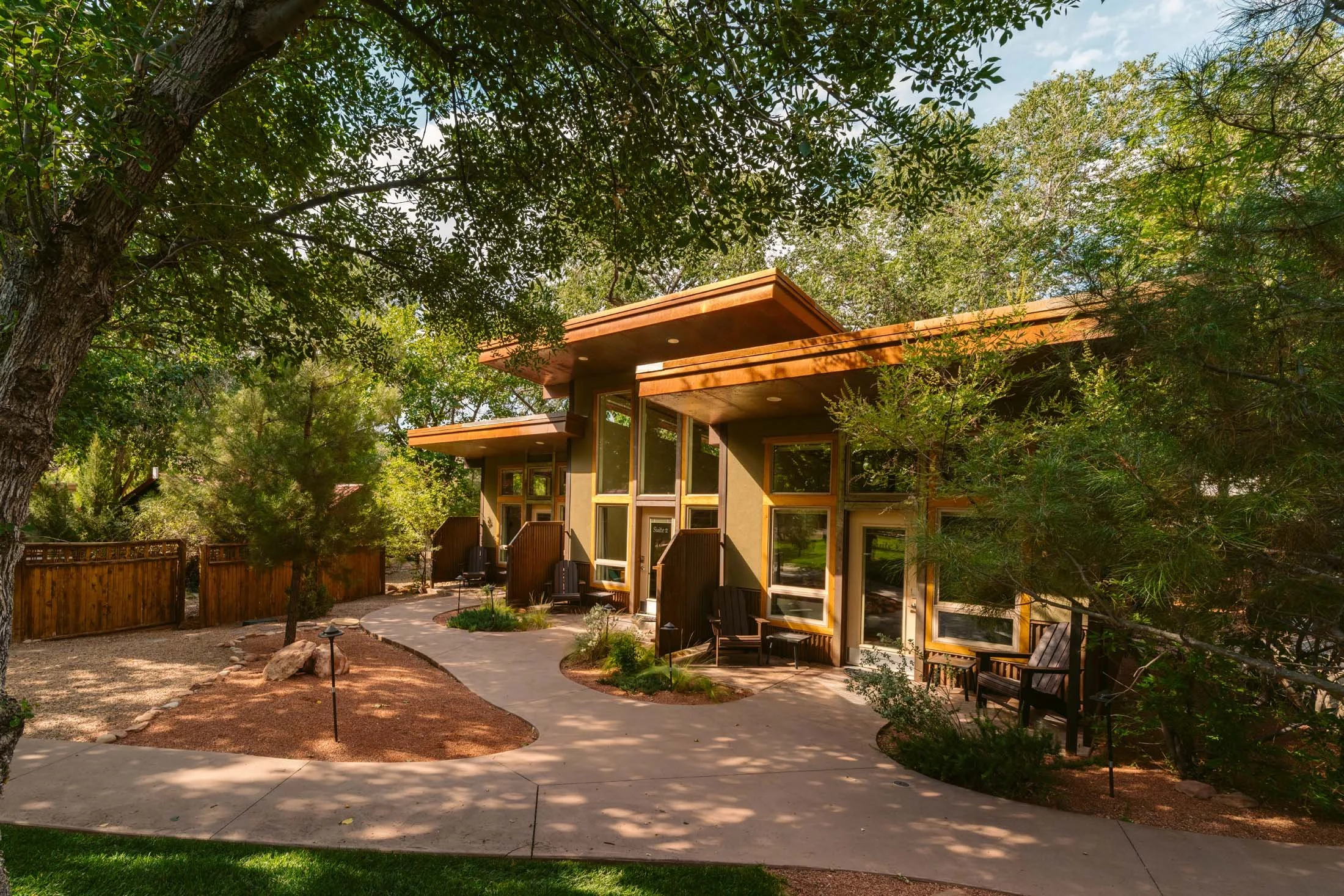 Modern multi-level house with large windows, wooden accents, surrounded by trees and a landscaped yard with a curved pathway and benches.
