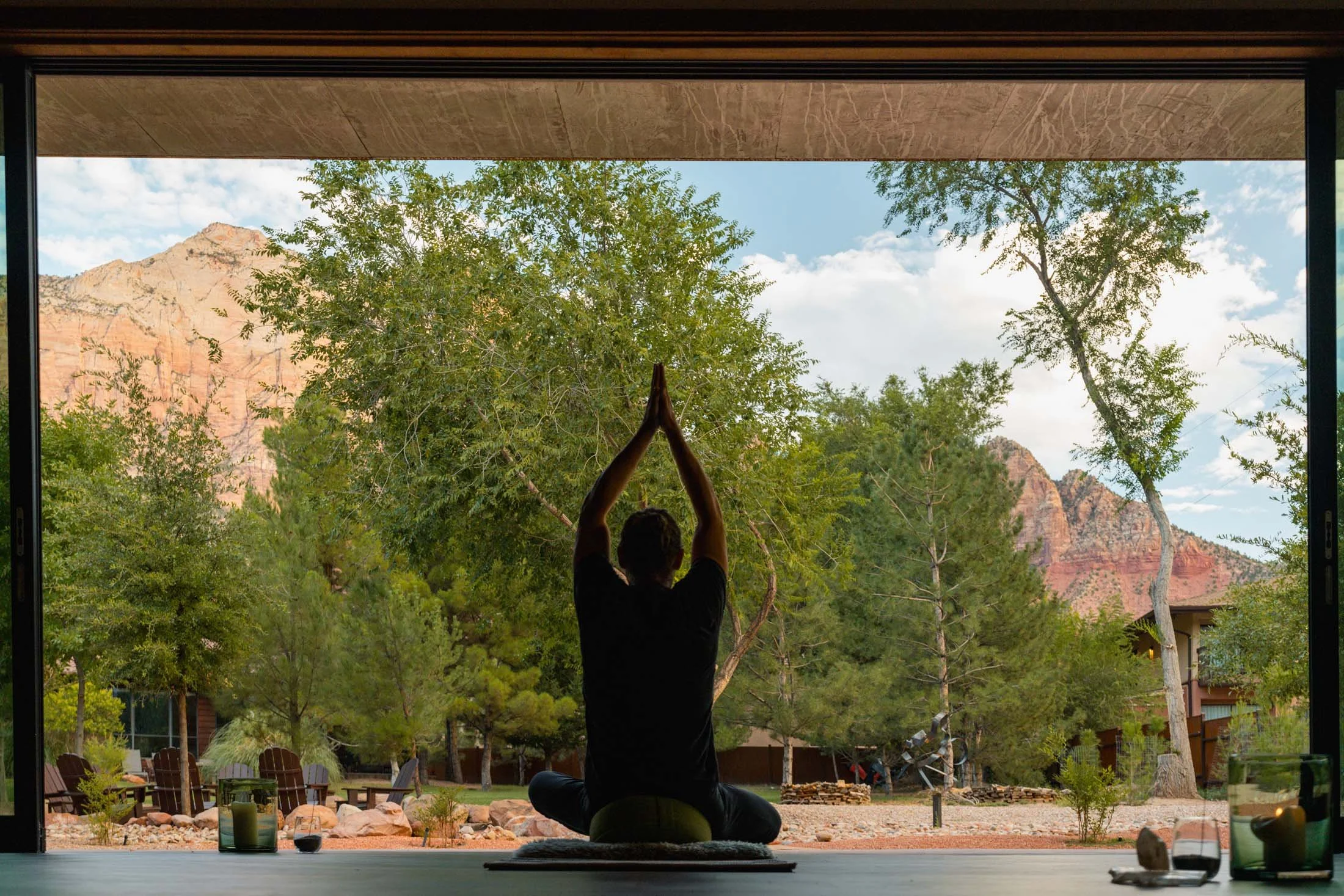 Person practicing yoga in a seated meditation pose with hands raised overhead, sitting on a mat indoors, looking out through large open windows to a scenic outdoor landscape with trees and mountains at Nama-Stay.