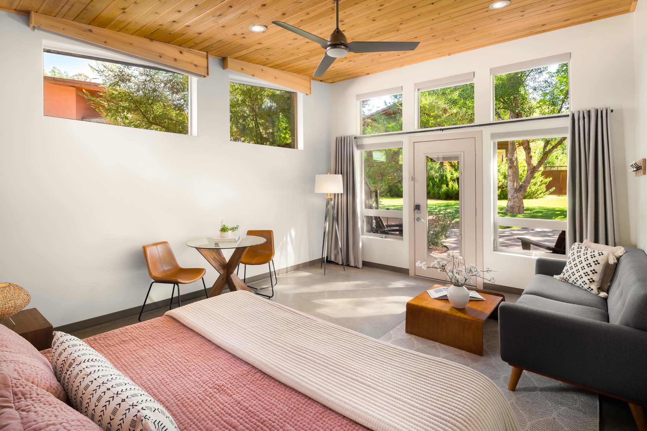 Living room with large windows, a ceiling fan, a gray sofa, a small coffee table with a flower vase, a floor lamp, a small round table with two chairs, and a view of trees outdoors.