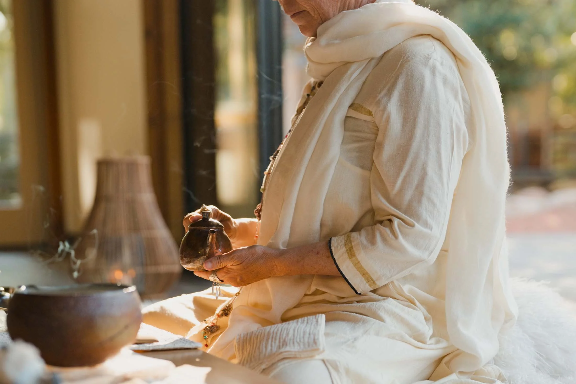 A woman dressed in traditional Indian attire, holding a small brass teapot, sitting indoors near a large window with sunlight streaming in.