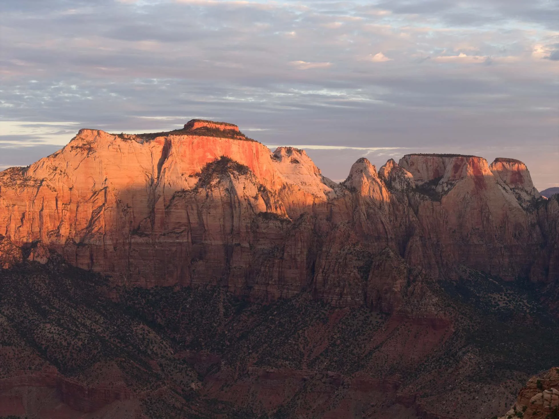 Sunset over a mountain range with reddish and beige cliffs and a partly cloudy sky.