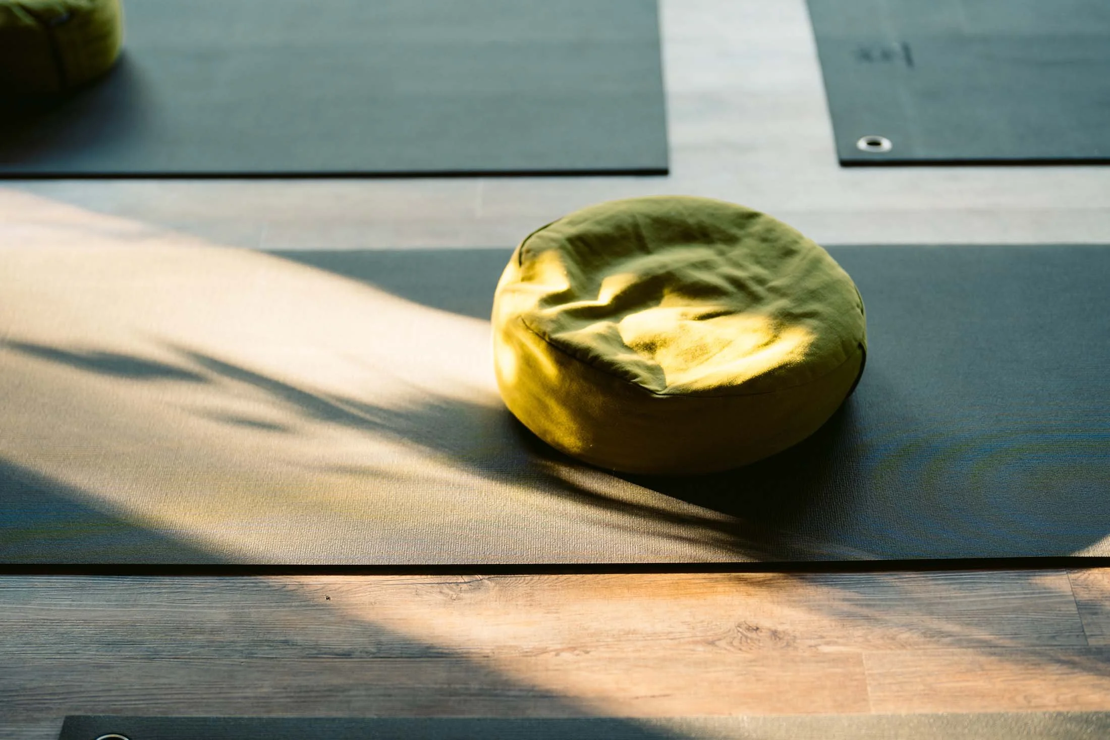 A yellow meditation cushion on a black yoga mat with sunlight and shadows cast on it, on a wooden floor.
