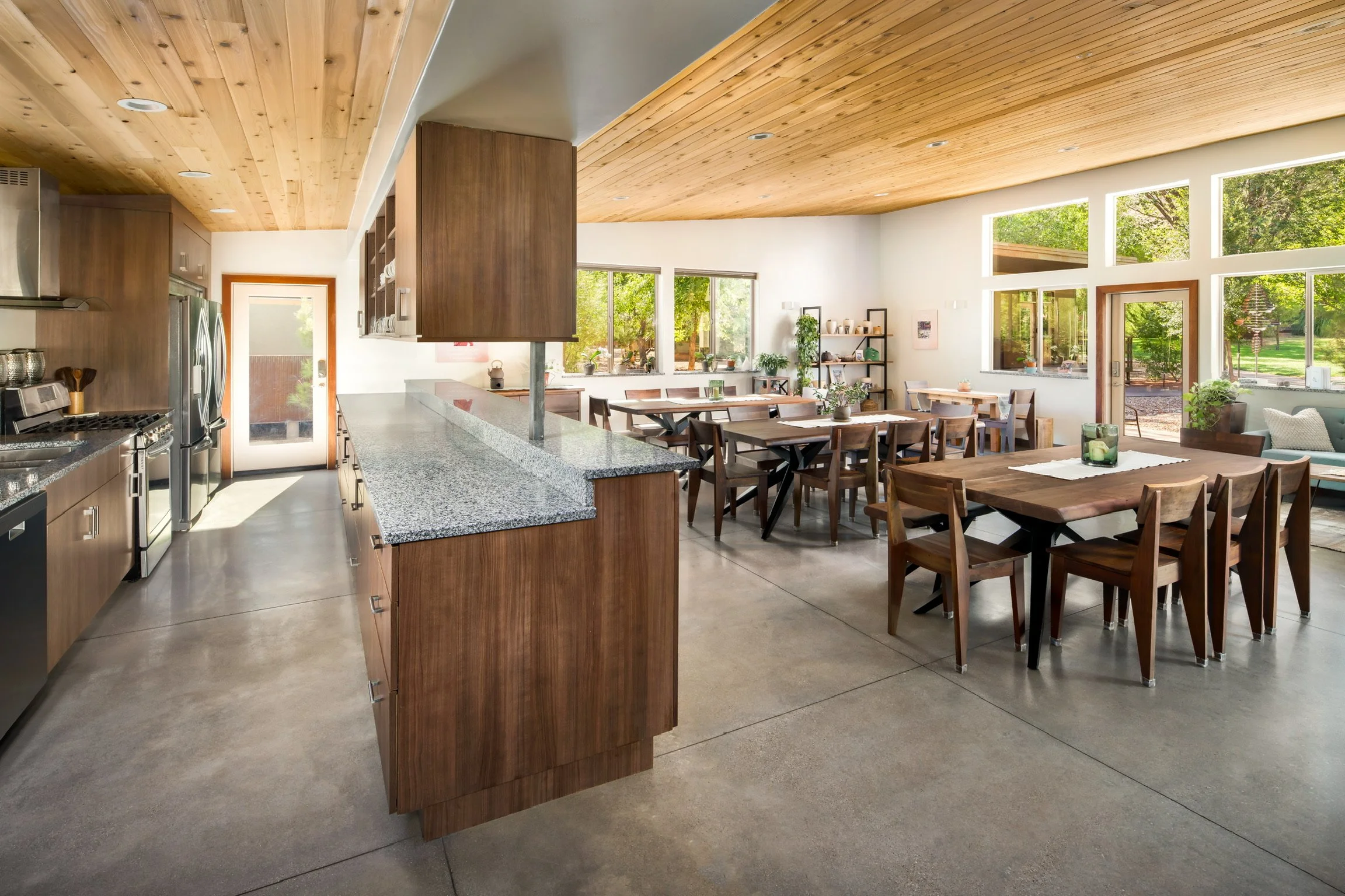 Spacious open-concept kitchen and dining area with wooden ceiling and large windows, featuring a granite kitchen island, wooden dining tables and chairs, and a door leading outside.